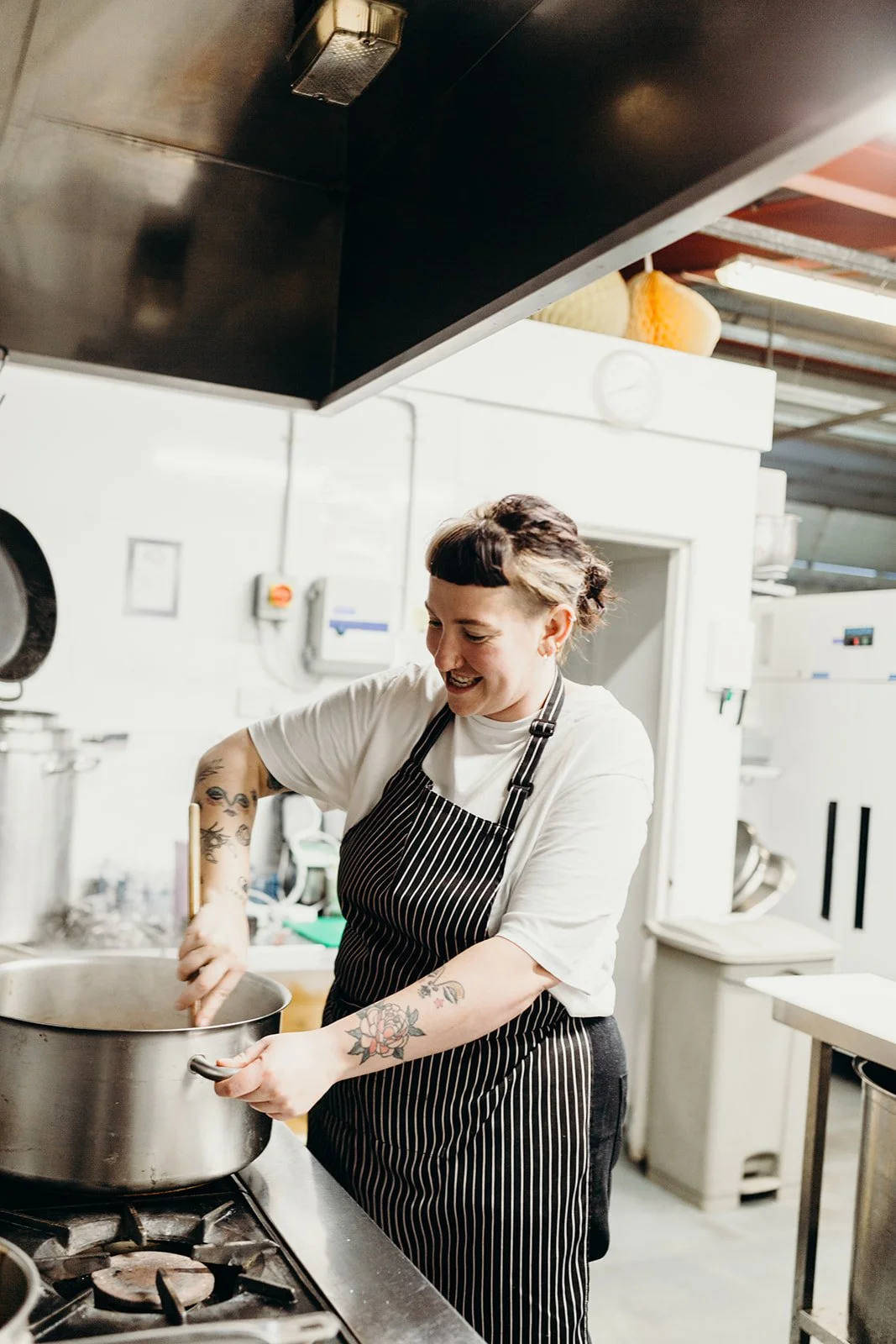 A woman with tattoos wearing a striped apron and white t-shirt stirring a pot in a commercial kitchen.