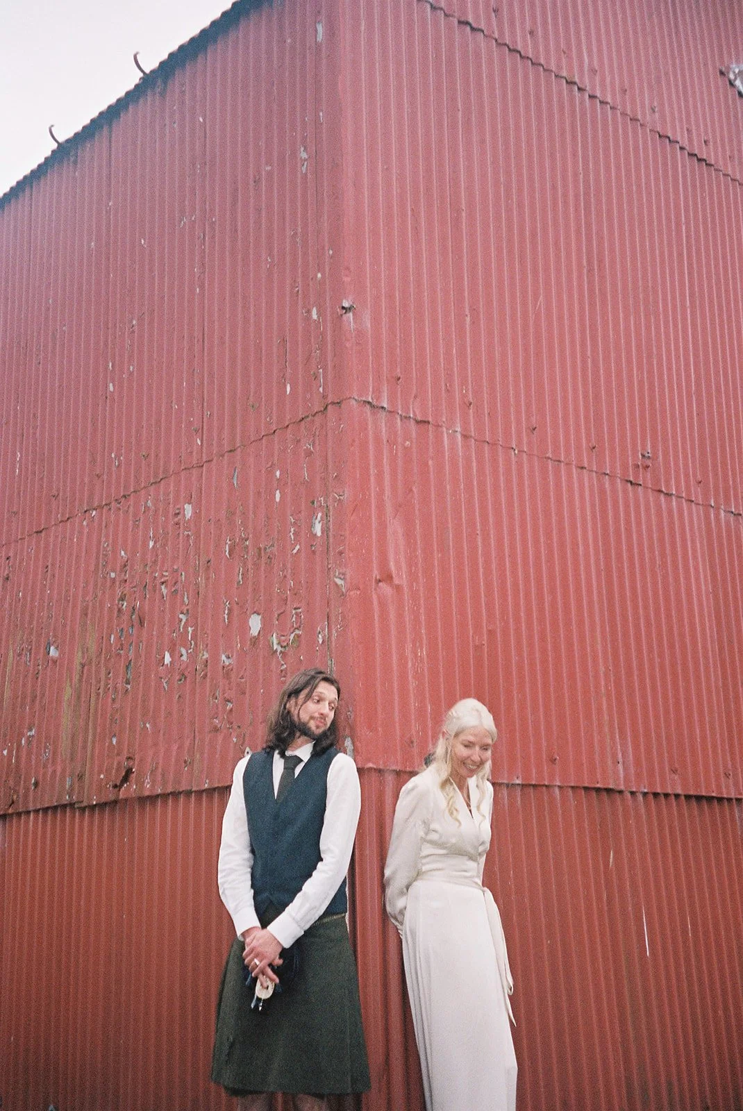 A man and woman standing in front of a large, red, weathered barn. The man has long hair, a beard, and is wearing a black vest, white shirt, black tie, and a long skirt. The woman has long blonde hair and is smiling, wearing a cream-colored dress.