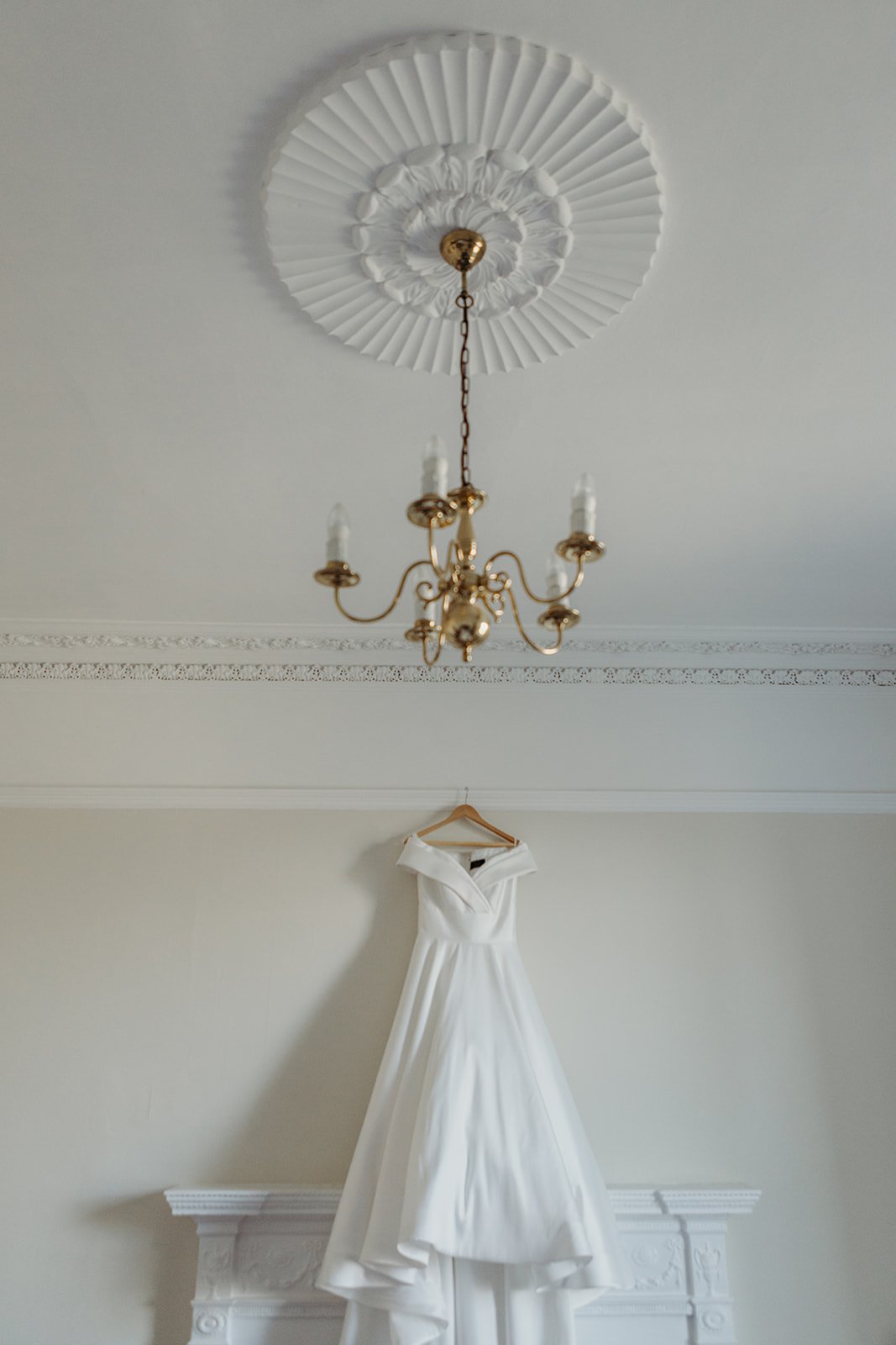 A white wedding dress hanging from a wooden hanger on a decorative mantelpiece in a room with intricate crown molding, a gold chandelier hanging from a decorative ceiling medallion.