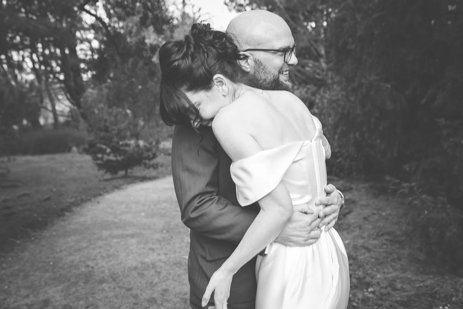 A couple embracing outdoors, the woman smiling with her head on the man's shoulder, in a park with trees in the background, black and white photograph.