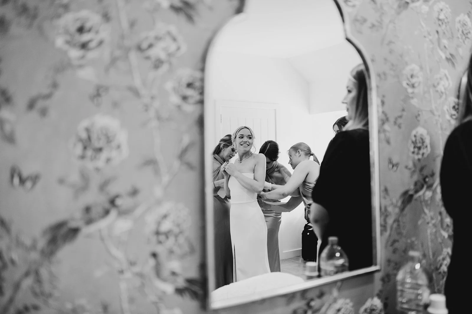 A woman in a wedding dress is smiling as she is helped by several women in a room, reflected in a mirror with floral wallpaper, preparing for a wedding.