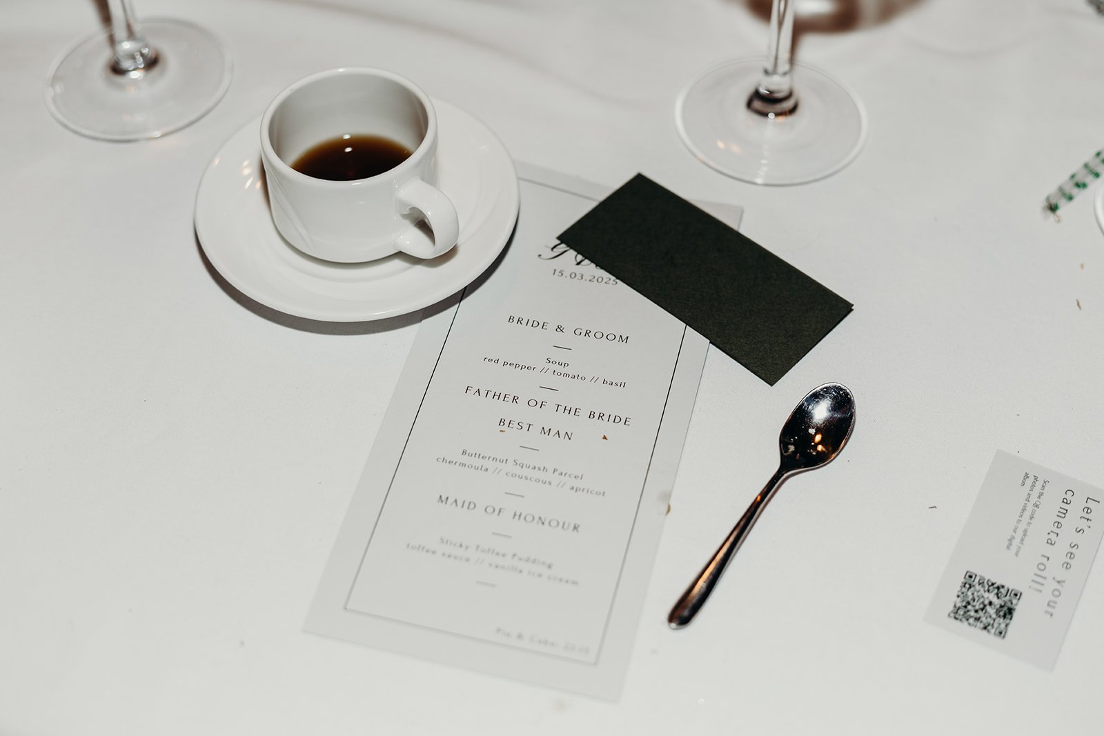 Table setting with wedding menu, black place card, cup of coffee on a saucer, silver spoon, and two empty wine glasses