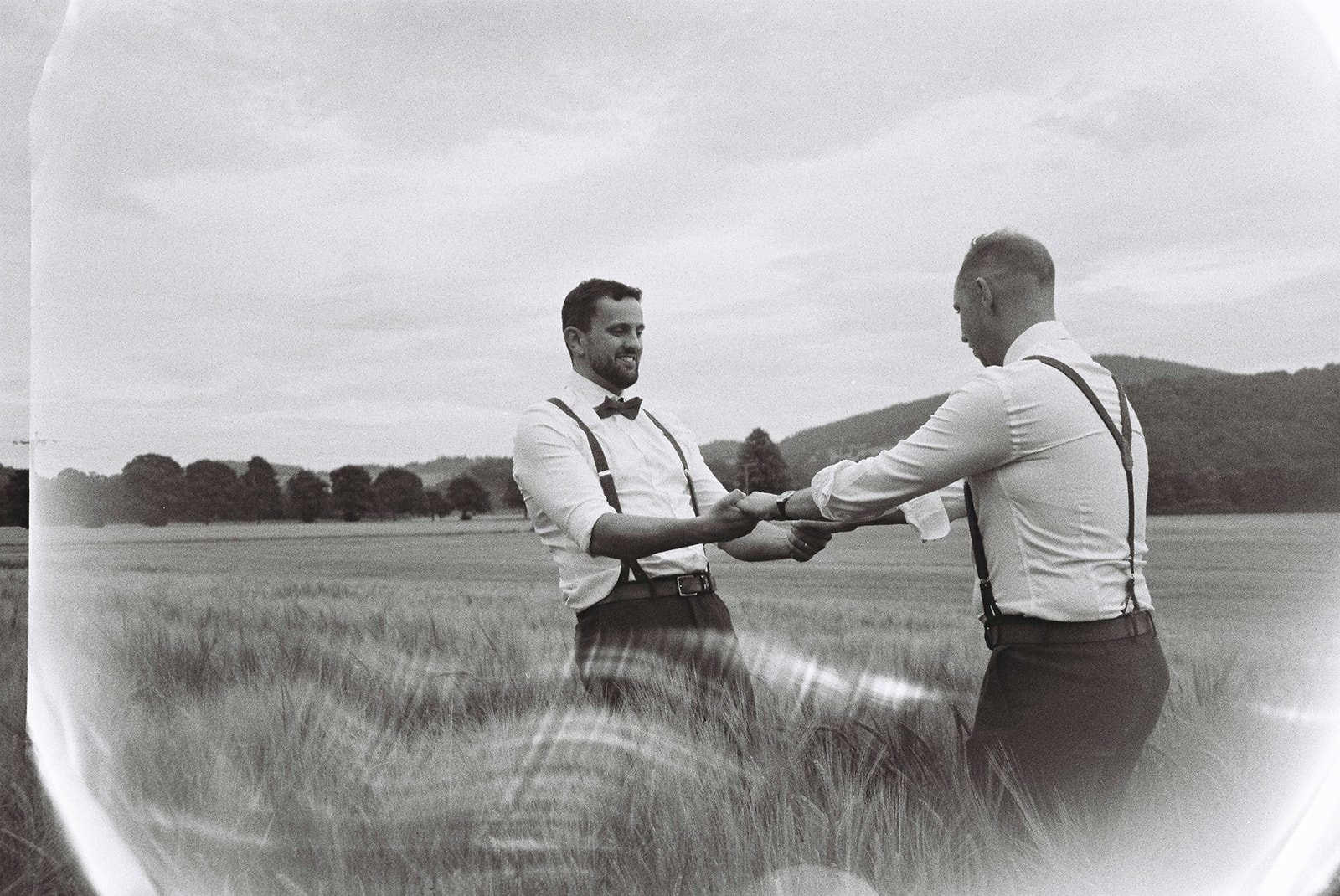 Two men in formal attire holding hands in a field with mountains in the background, smiling at each other.