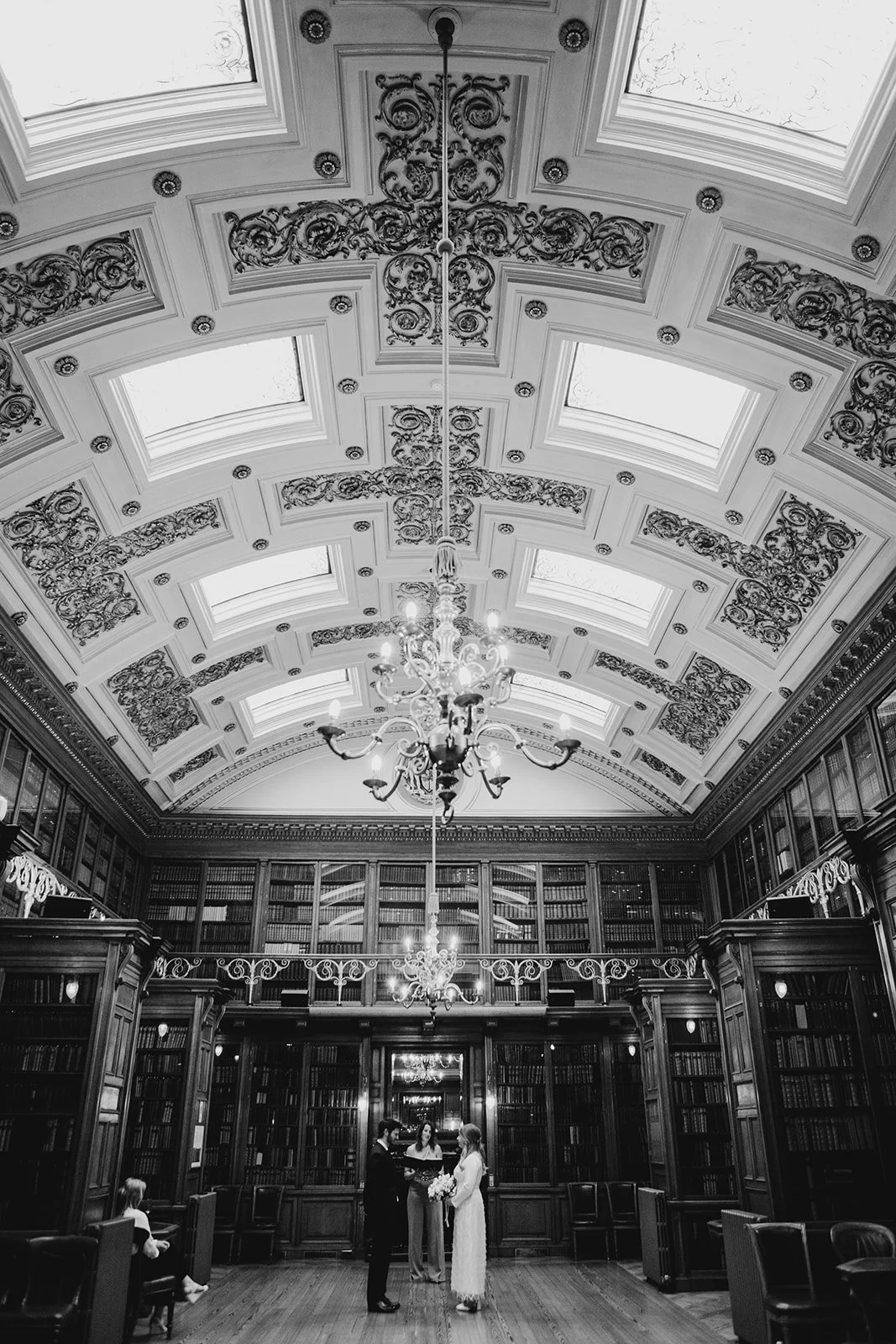 A wedding ceremony taking place in an elegant, historic library with tall bookshelves and ornate ceilings, featuring a chandelier and two brides and a groom standing in front of an officiant.