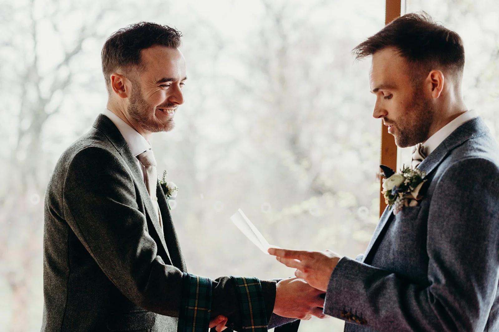 Two men in suits holding hands during a wedding ceremony, exchanging vows indoors with a window and natural light behind them.