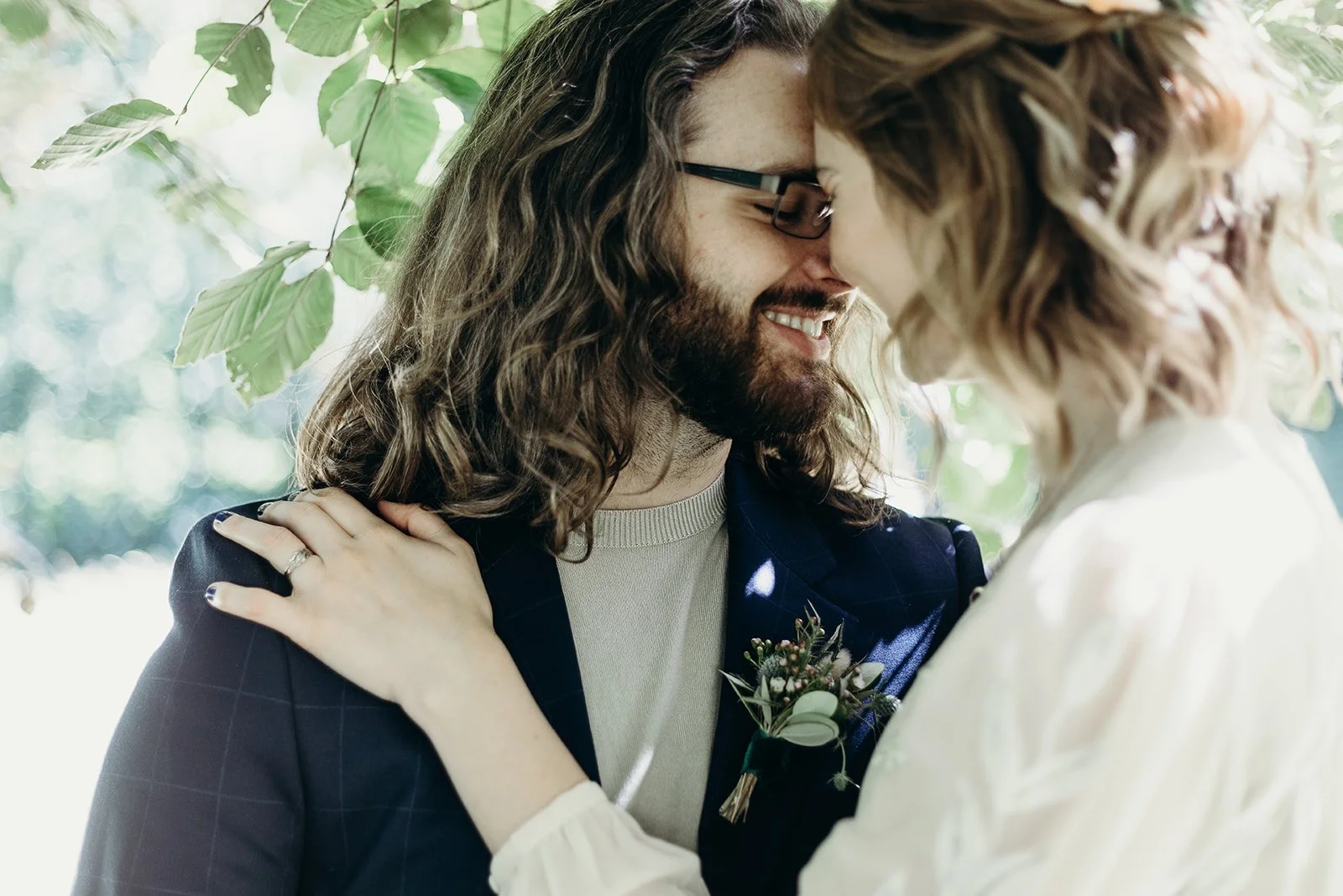 A couple with long hair and glasses, smiling and touching foreheads, surrounded by green leaves.