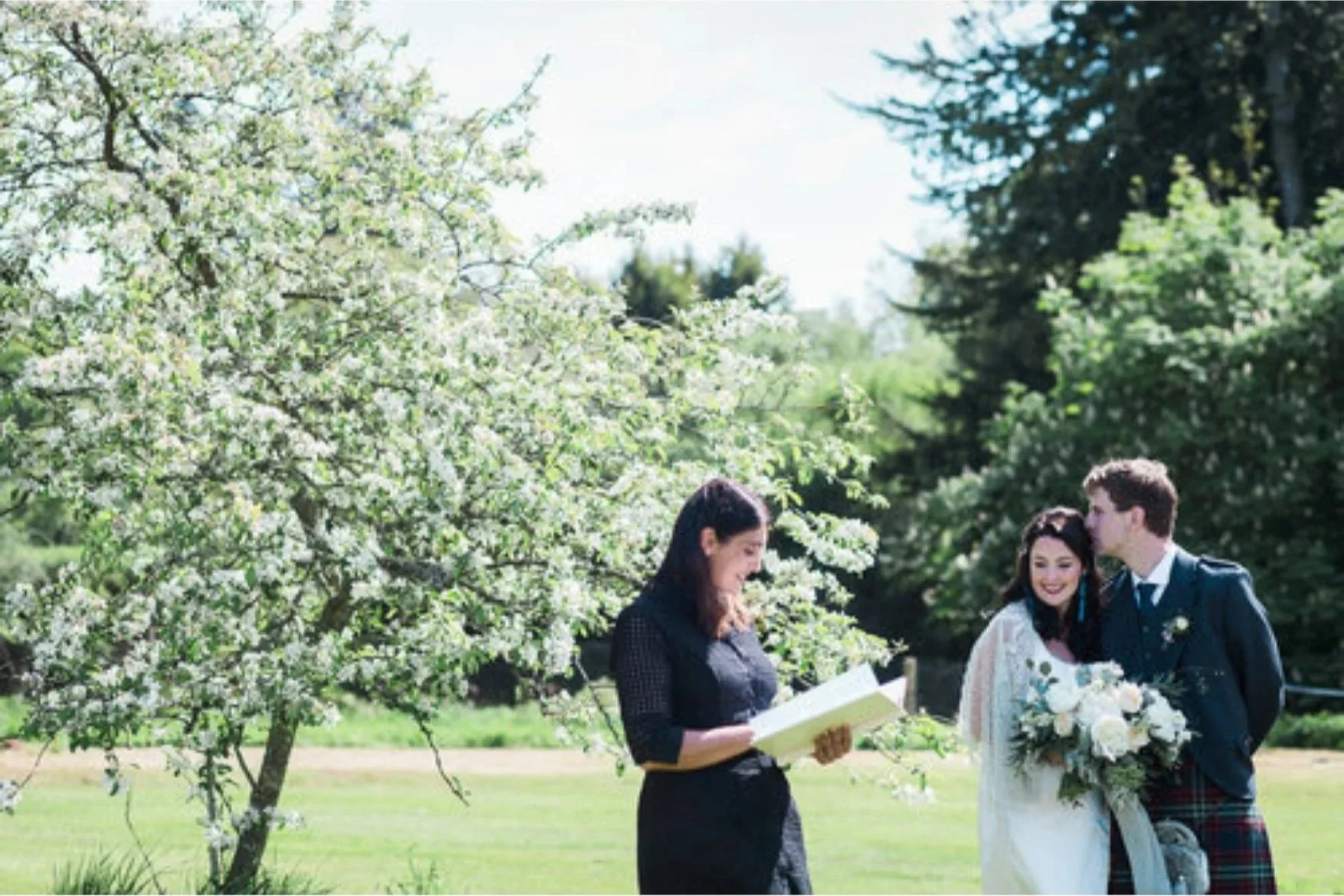 A wedding ceremony outdoors with a bride and groom standing together, holding flowers, and a woman reading from a book under blooming trees on a sunny day.