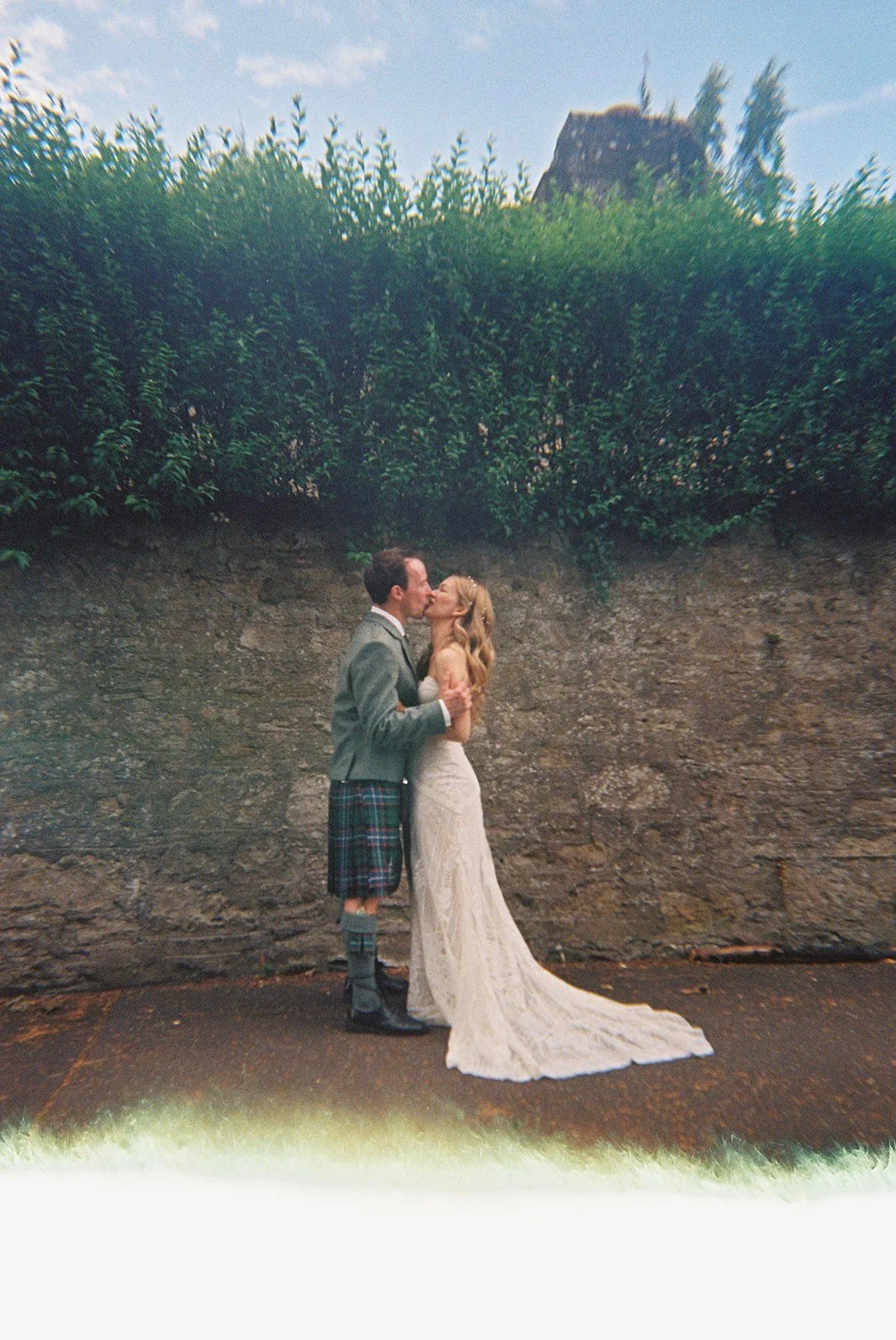 A bride and groom share a kiss outdoors in front of a stone wall with bushes, one of the groom's legs is covered in a kilt and knee-high socks, the bride is wearing a long white lace wedding dress.