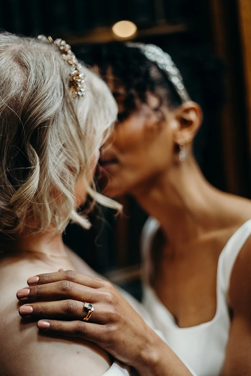 Two women, one with blonde curly hair and the other with short dark hair, sharing an intimate moment, wearing jewelry and strapless white dresses.