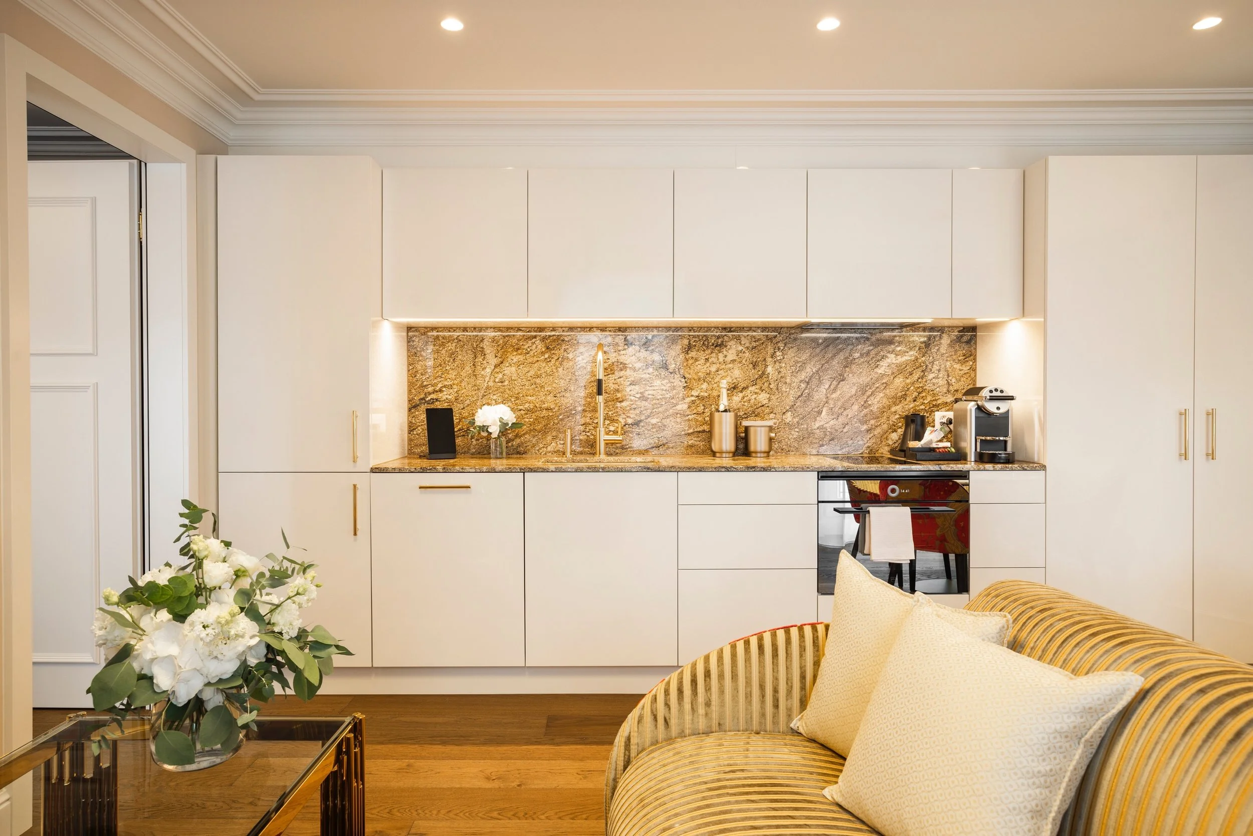 Hotel apartment kitchen and sitting area with light cabinetry and patterned furnishings.