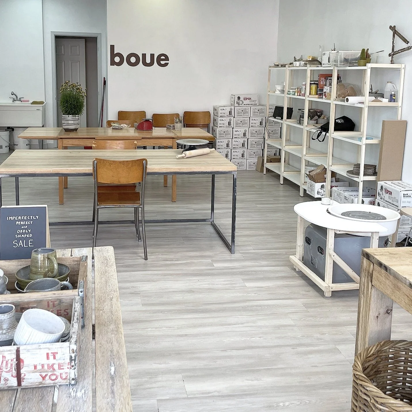 Interior of a pottery studio with tables, chairs, shelves with supplies, and a pottery wheel. A sign on the wall reads "boue." Boxes and ceramics are displayed throughout the space.