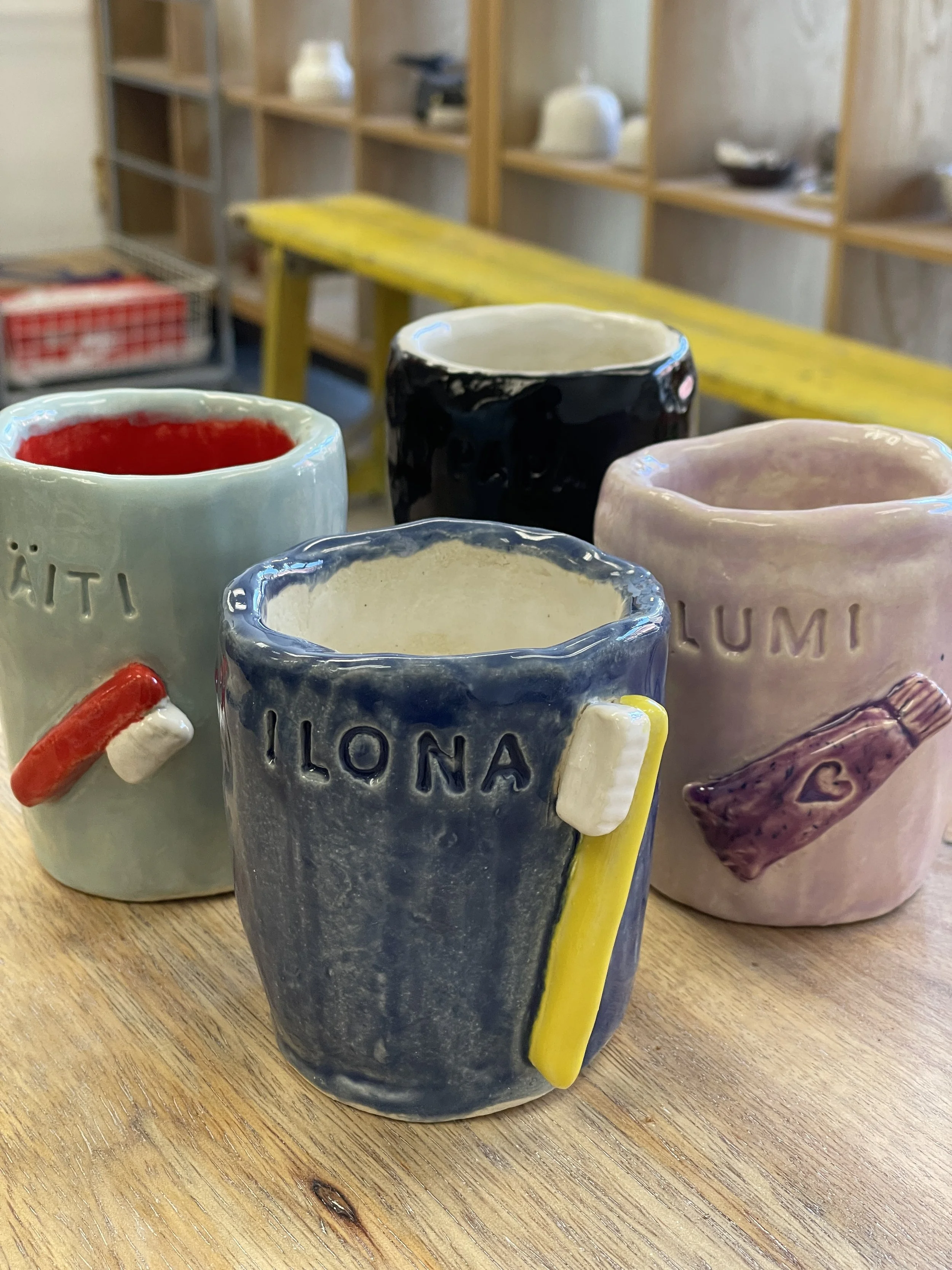 Four colorful ceramic mugs on a wooden table, each adorned with small 3D sculpted toothbrushes and toothpaste tubes. Background shelves are visible.