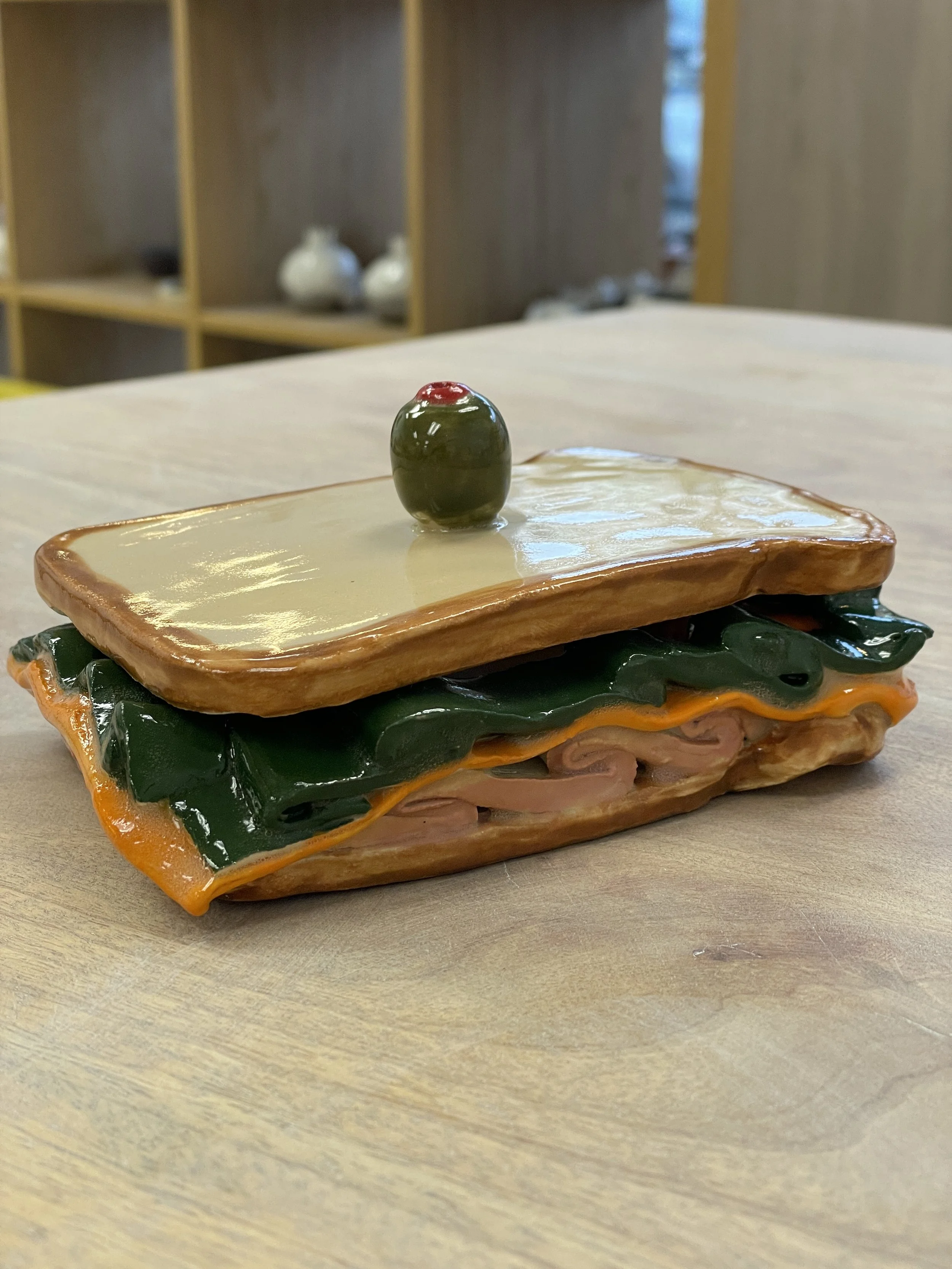 Ceramic sculpture of a sandwich on a wooden surface. The sandwich features layers of meats, cheese, and lettuce, topped with a ceramic olive on a toothpick.