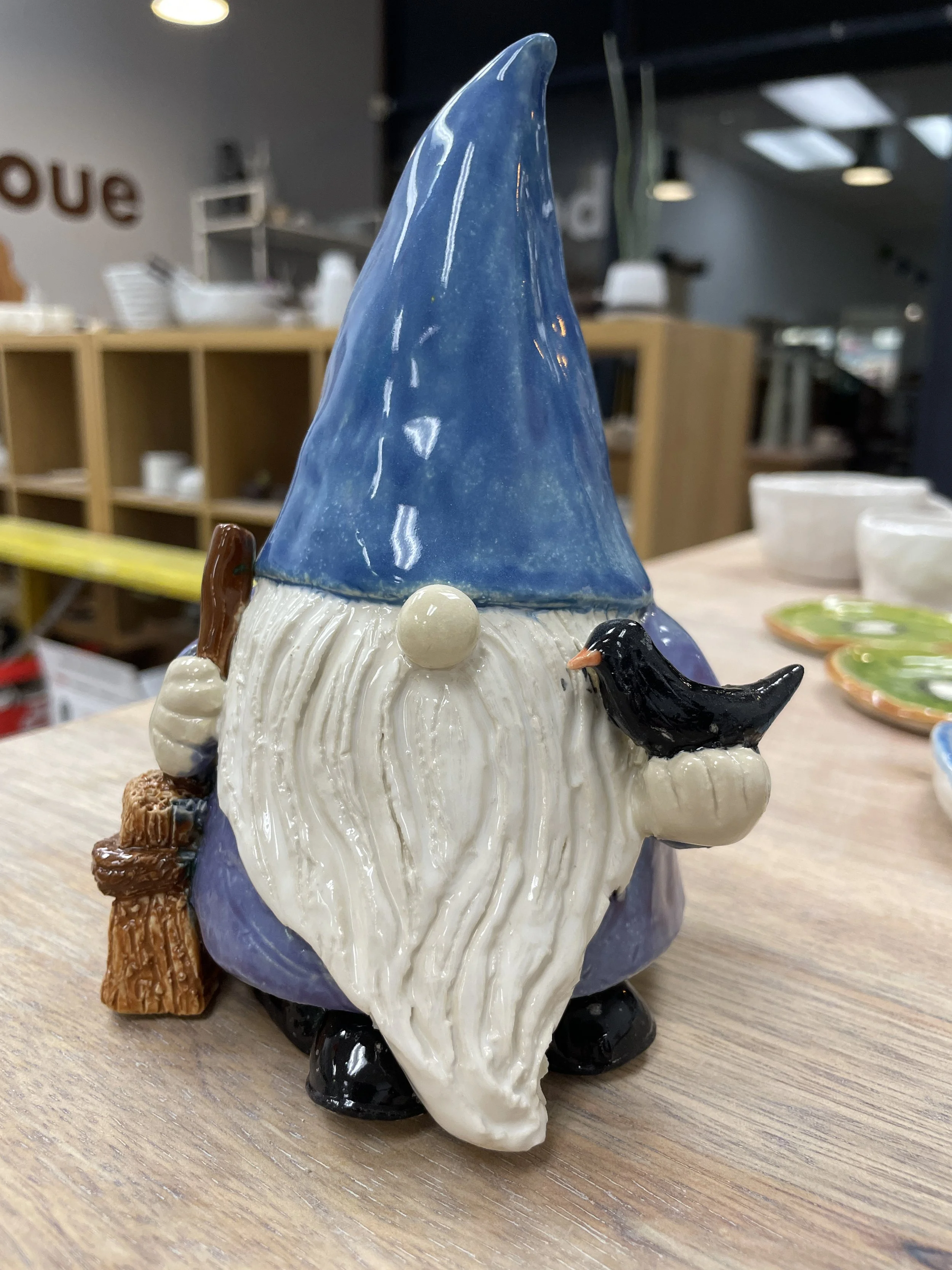 Ceramic gnome figurine with a blue hat, white beard, holding a black bird, and standing on a wooden stick, displayed indoors on a table.