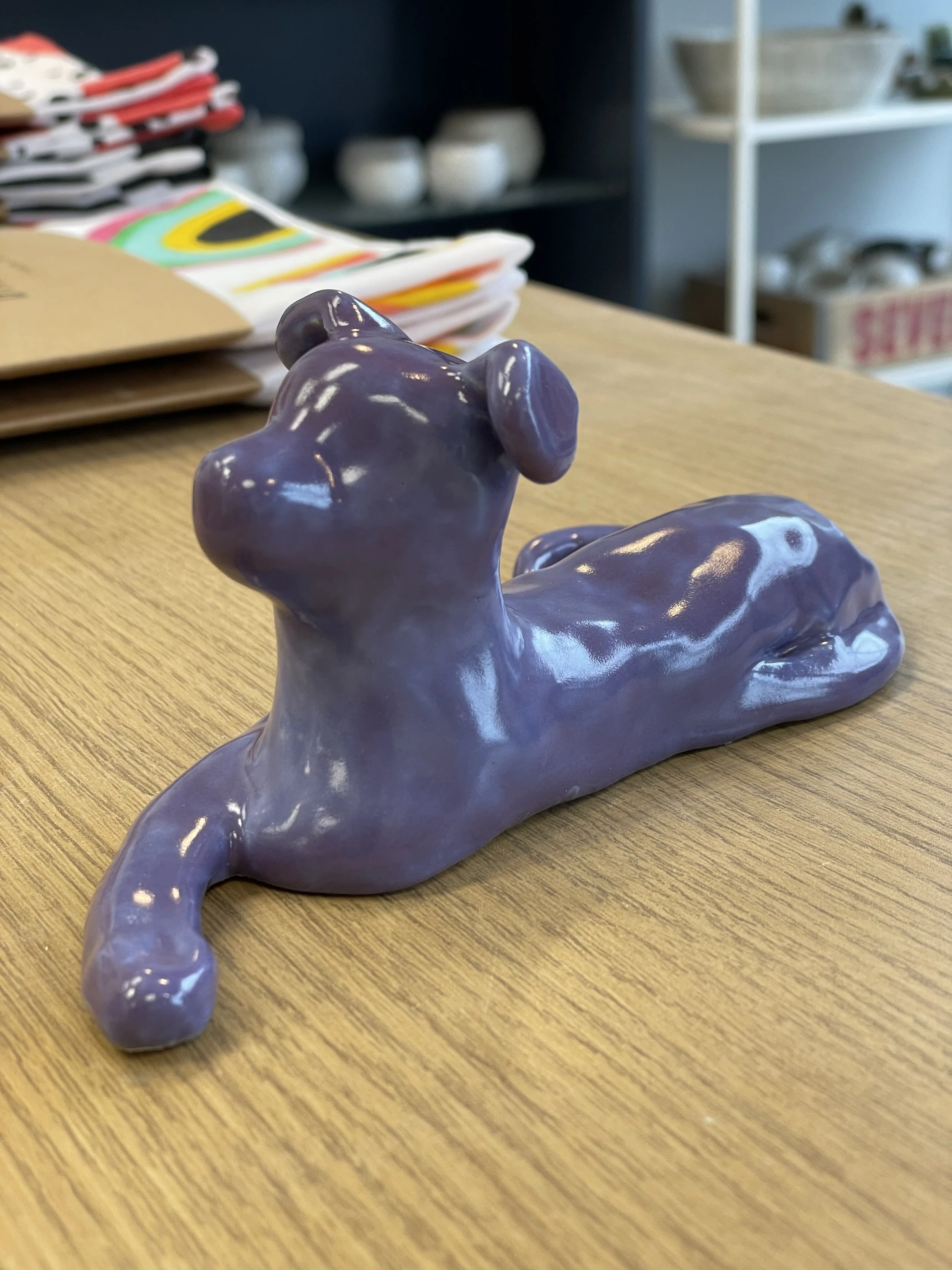 Purple ceramic dog sculpture on wooden table with background shelves.
