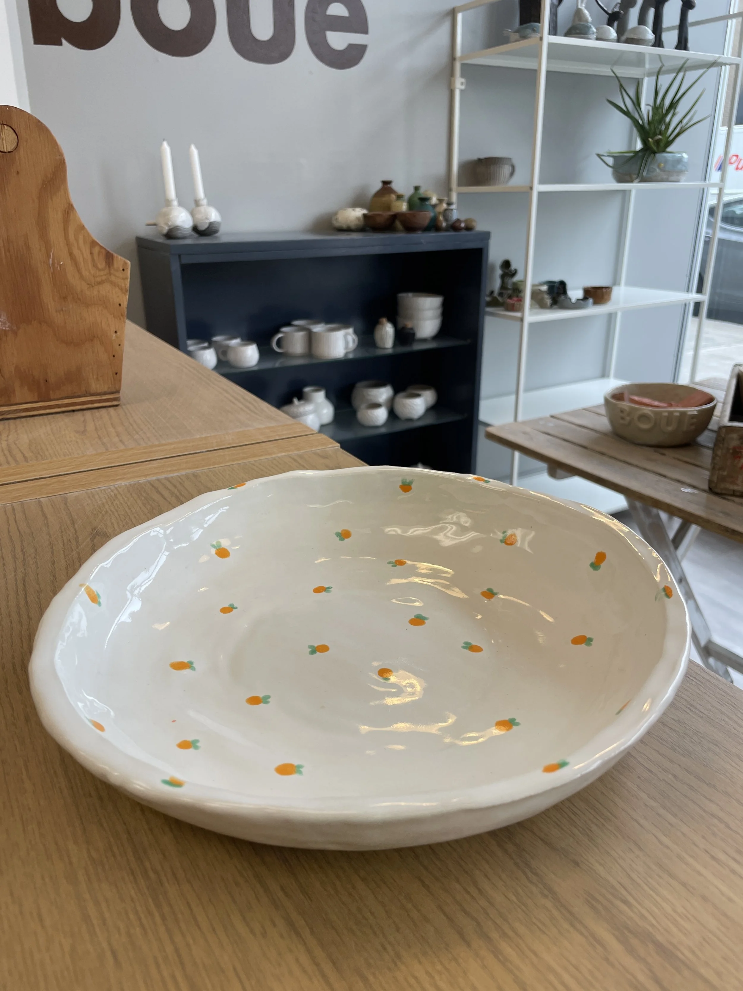 A white ceramic bowl with small orange and green patterns on a wooden table, in a shop with shelves displaying pottery and plants.