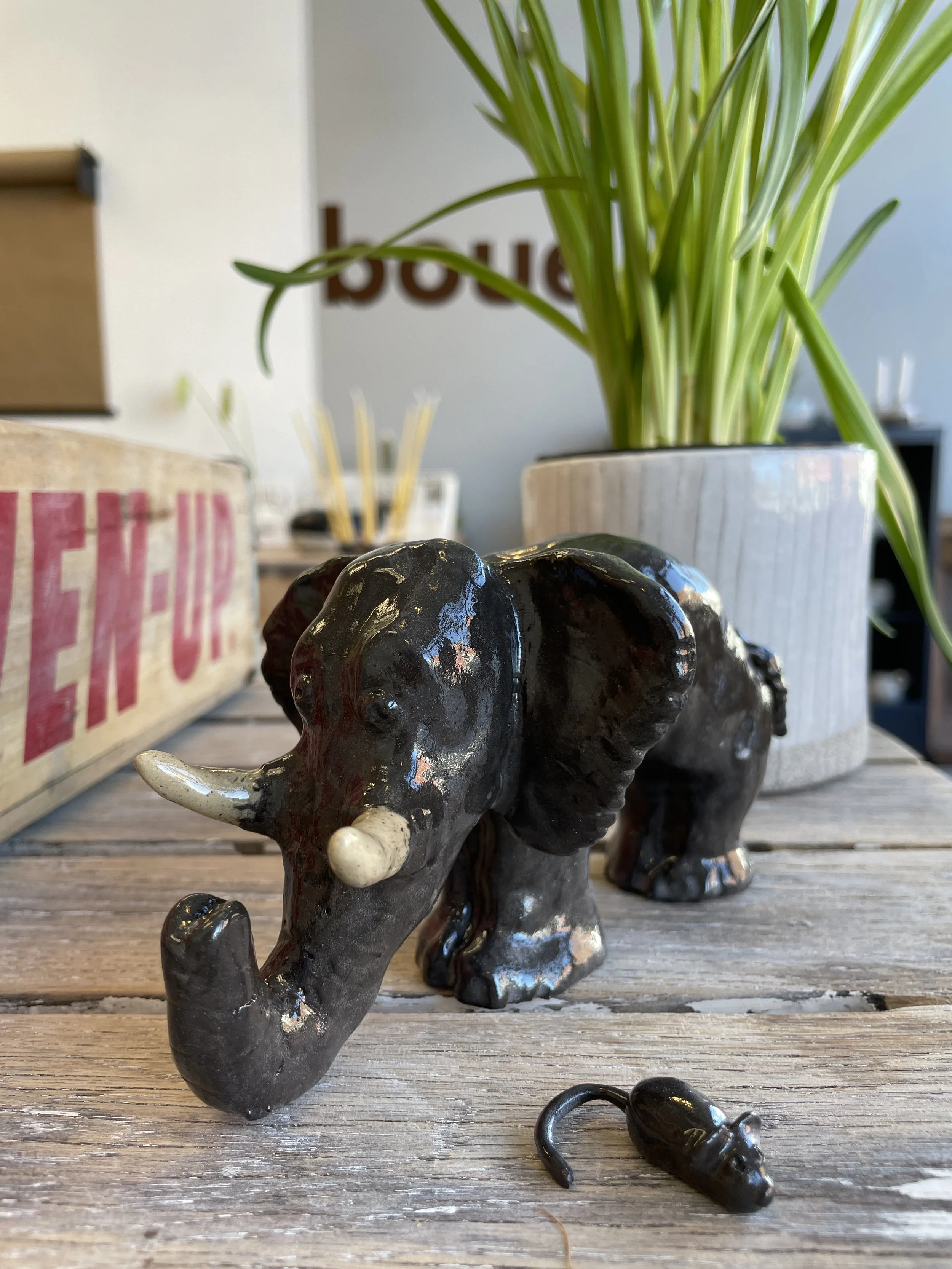 Black ceramic elephant figurine on a rustic wooden table, with a small ceramic mouse figurine nearby. A green plant in a white pot is in the background, along with a partially visible wooden crate.