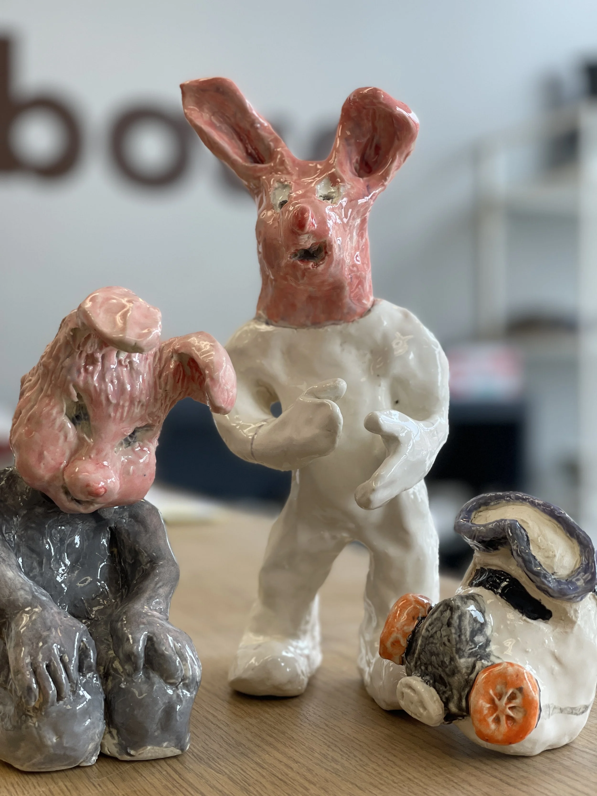 Three ceramic sculptures with rabbit-like features; one seated, one standing, and one abstract piece with orange and black elements. They have textured, glossy surfaces and are placed on a wooden surface.
