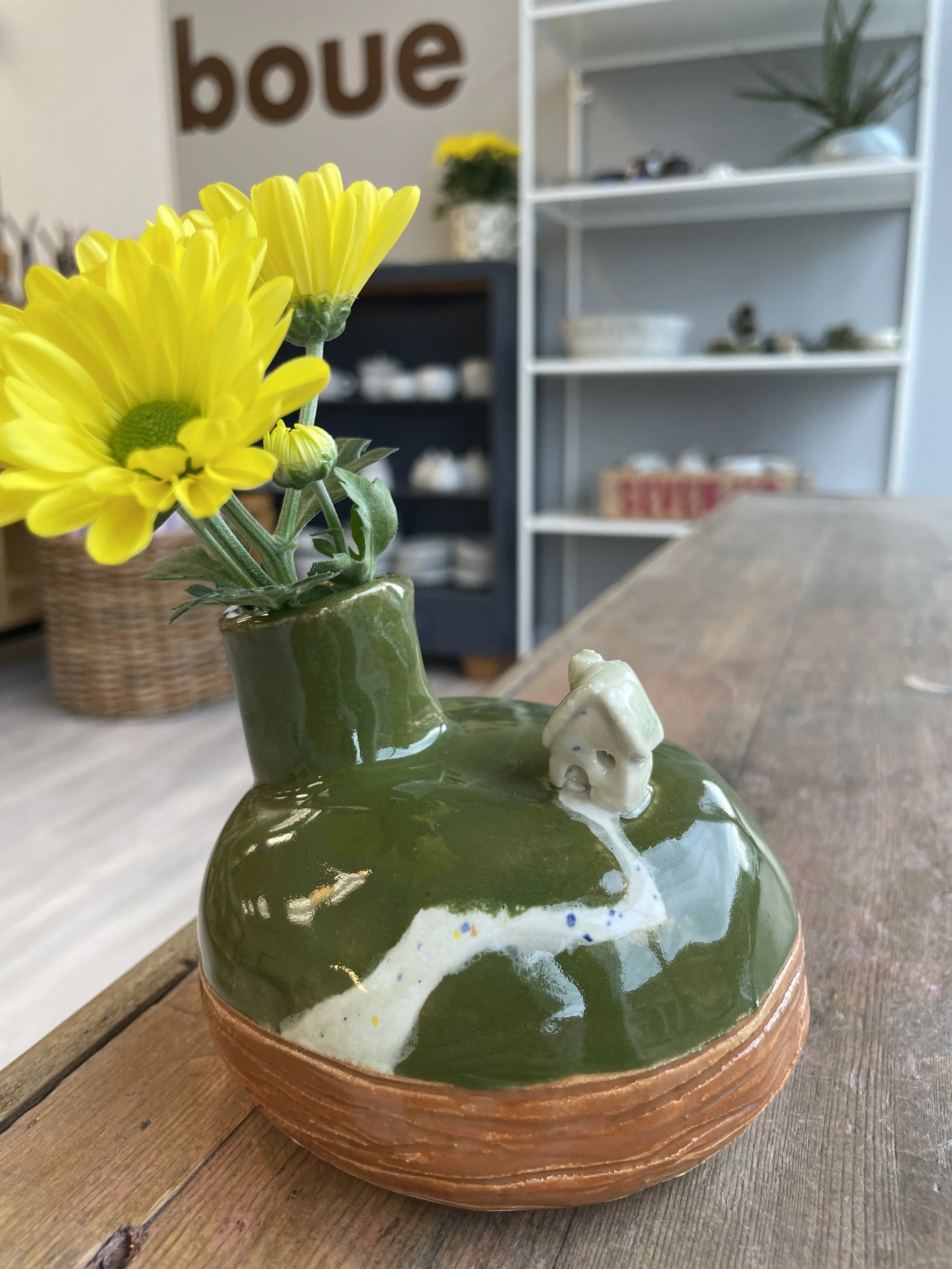 Small green ceramic vase with a miniature house and path design, holding yellow flowers, on a wooden table in a shop with shelves in the background.