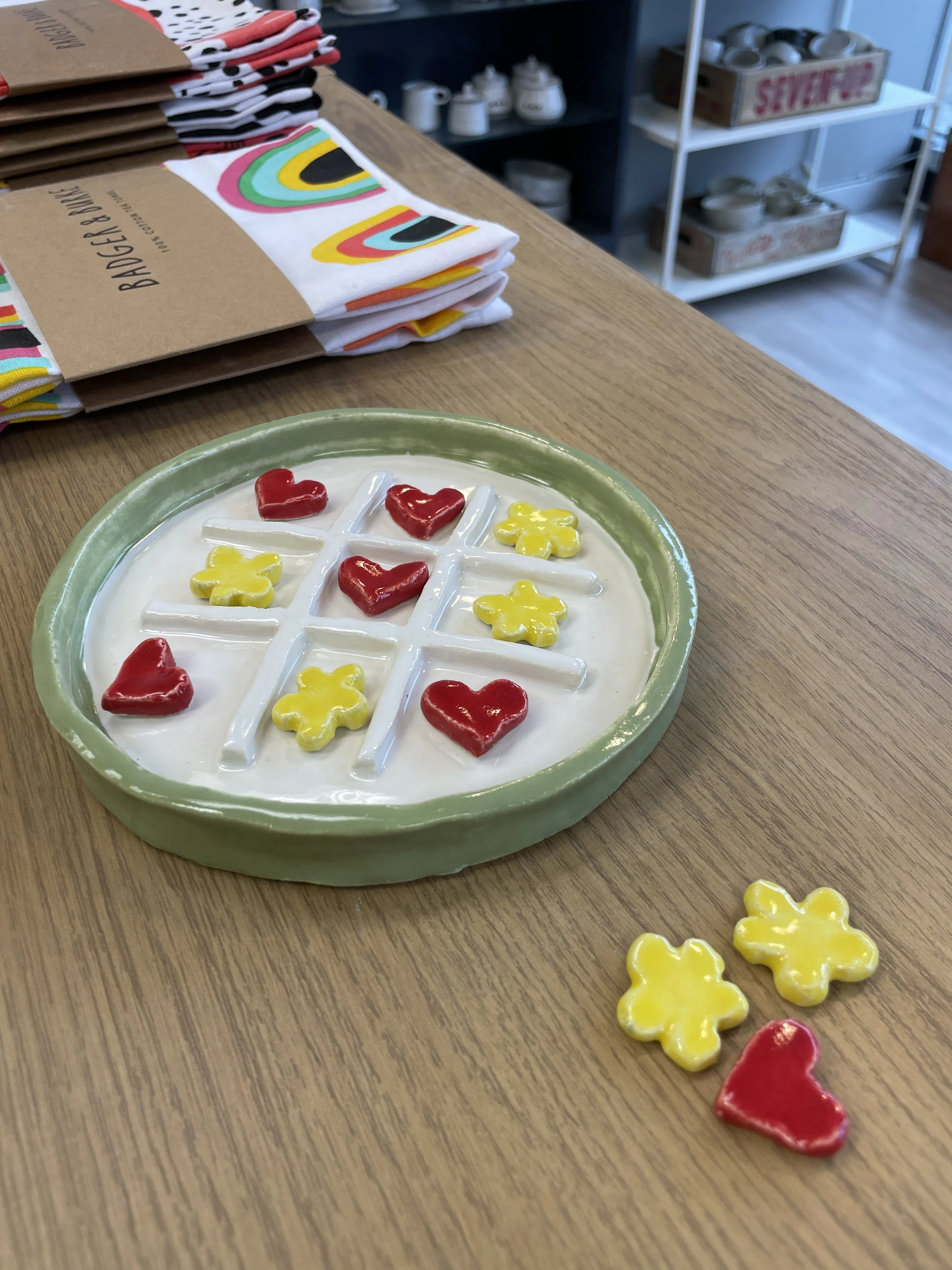 Ceramic tic-tac-toe set with red hearts and yellow flowers on a wooden table, with colorful folded towels in the background.