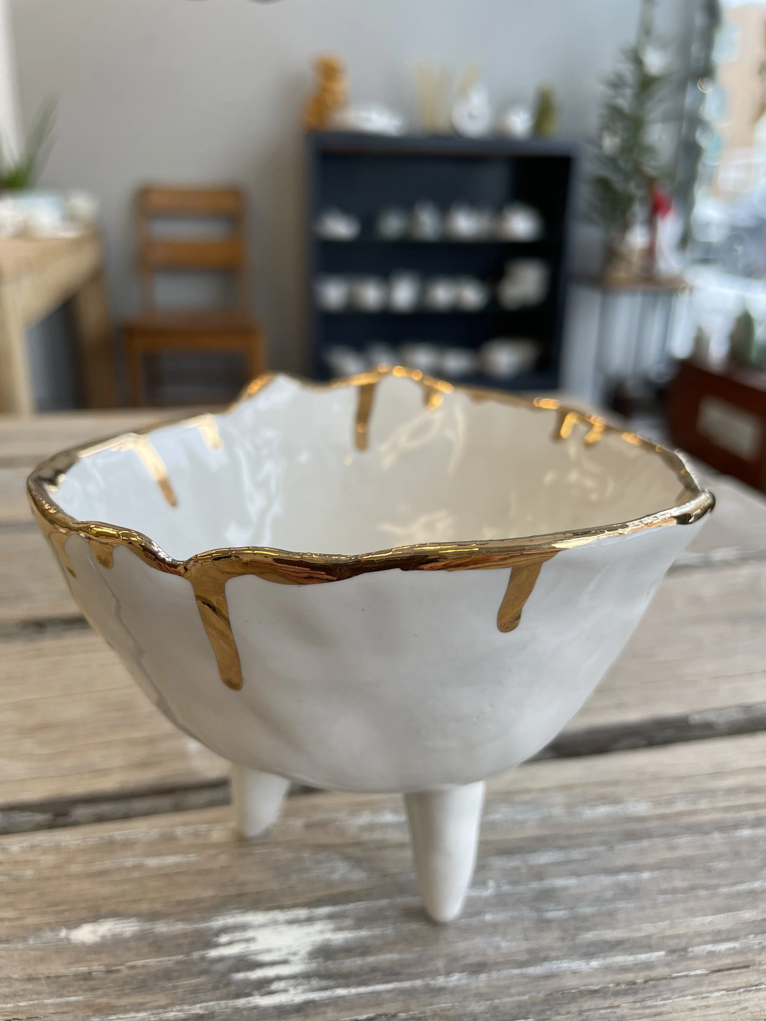 White ceramic bowl with gold accents on a wooden table, background shows a blurred interior with shelves and chairs.