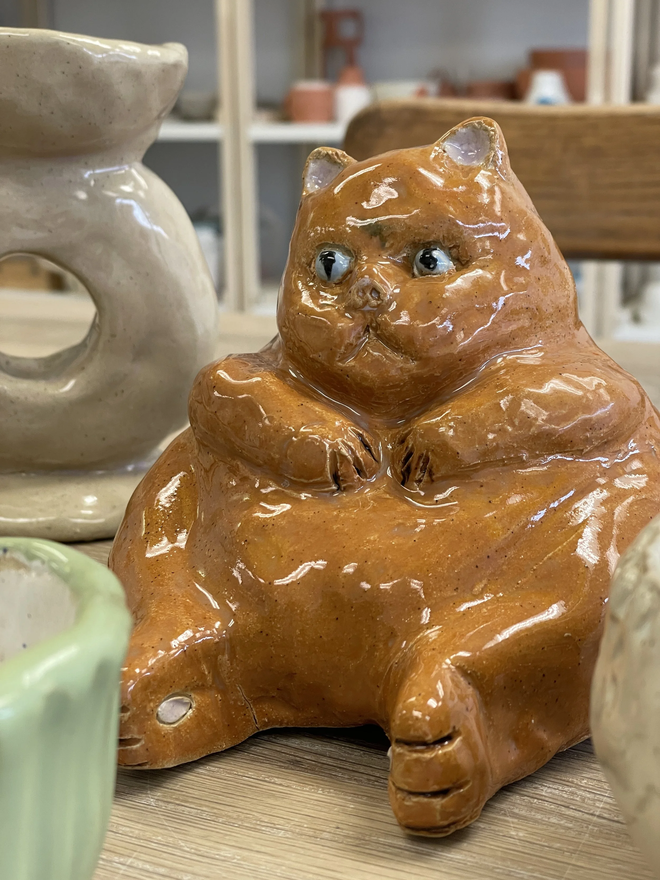 Ceramic sculpture of a plump cat with a glossy brown finish, sitting among other pottery on a wooden surface.