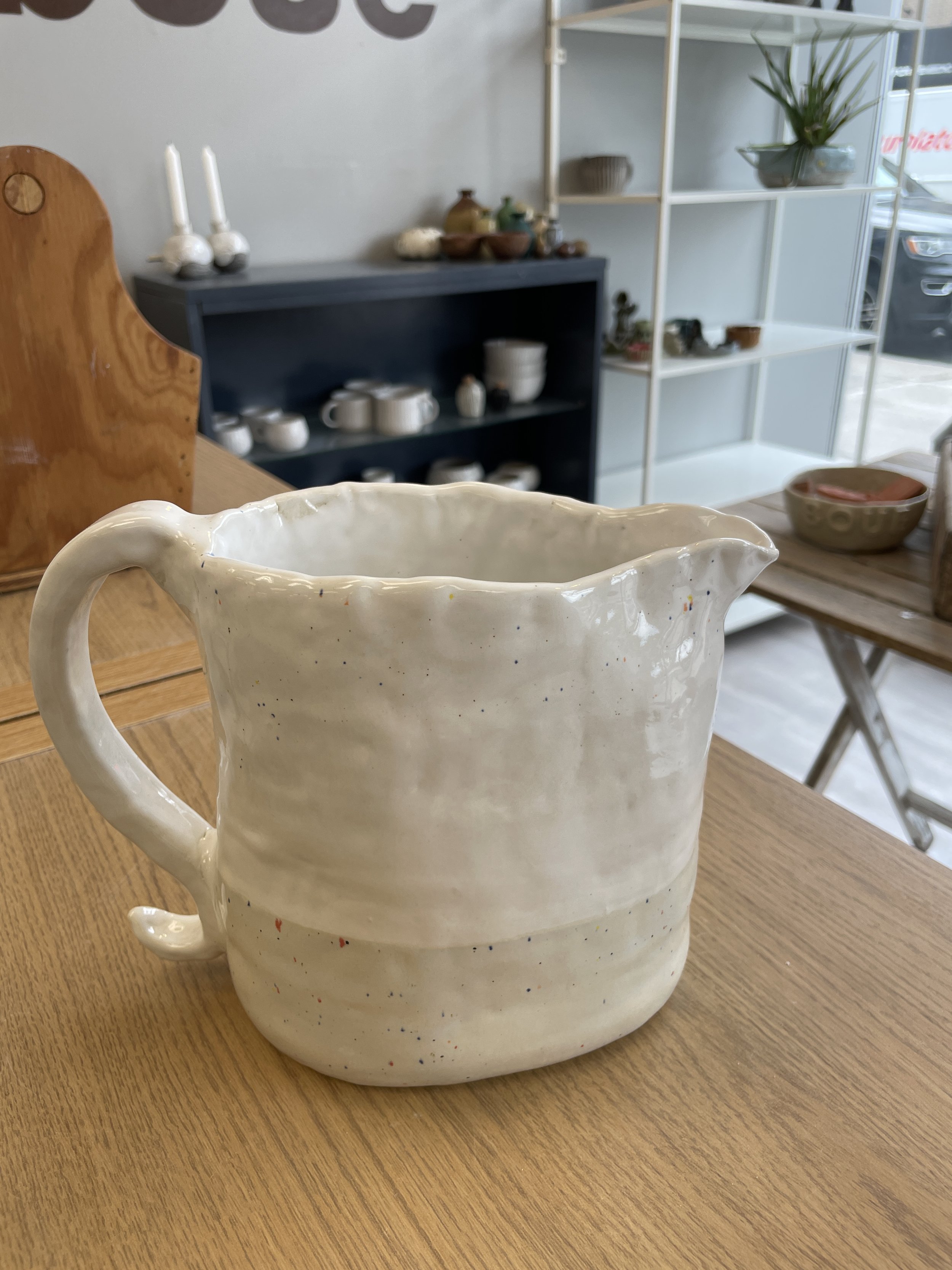 Ceramic pitcher on wooden table in pottery store, with shelves and pottery in background.