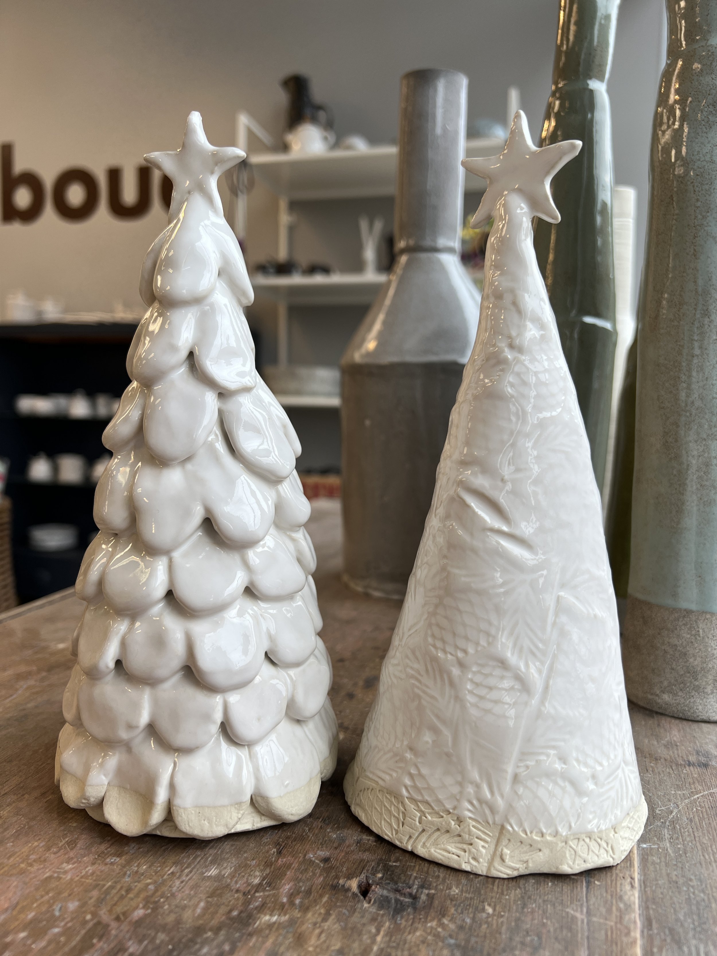 Two ceramic Christmas tree ornaments with star tops on a wooden surface in a shop.