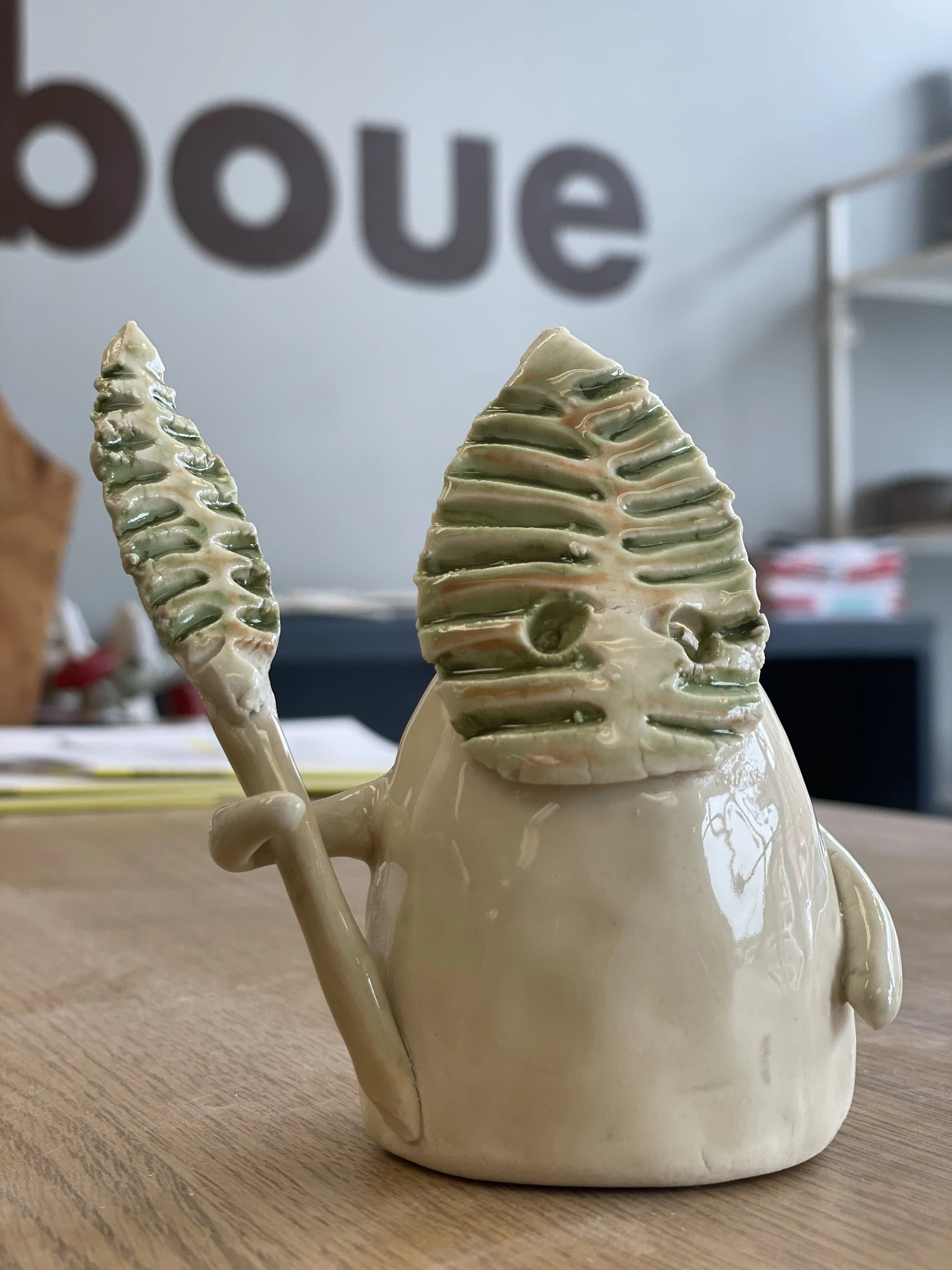 Ceramic sculpture of a small, abstract figure with a green leaf-patterned head holding a leafy staff, placed on a wooden table. Blurred background with partial text.