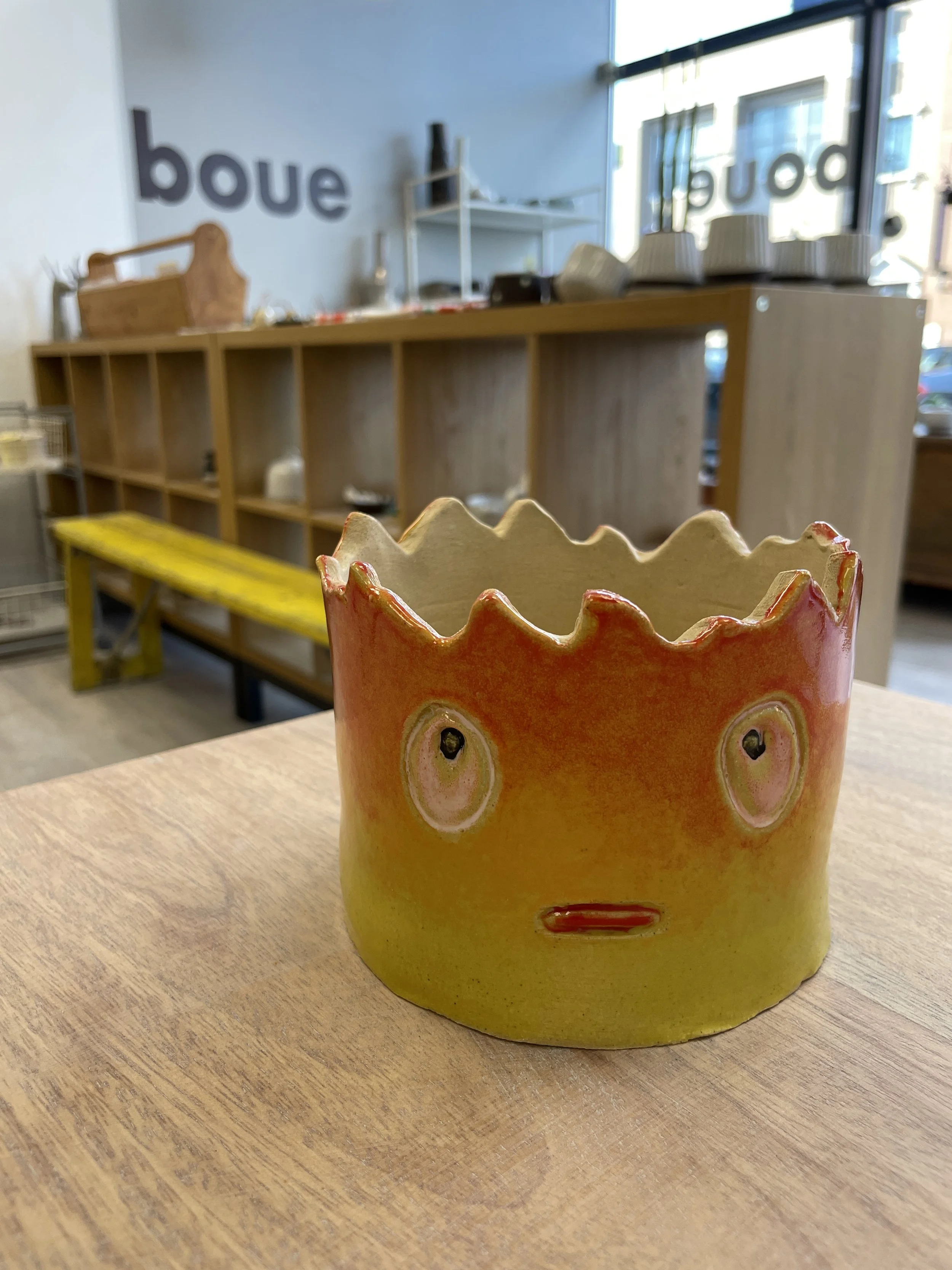 A ceramic pot with a face and jagged top on a wooden table in a store with shelves in the background.