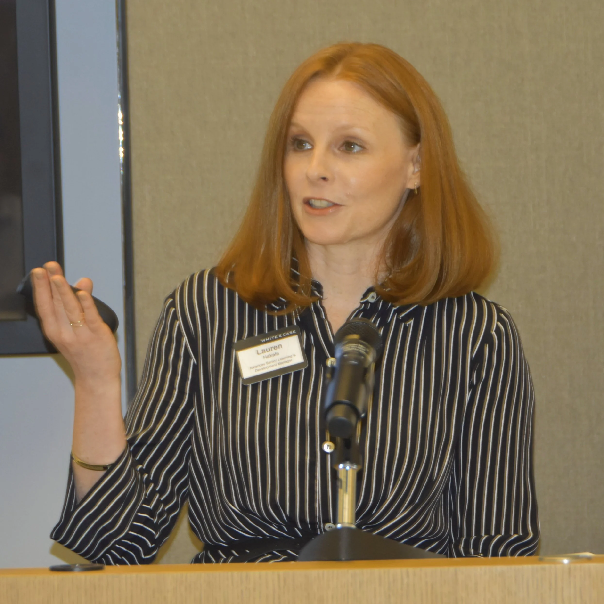 A woman with red hair wearing a black and white striped shirt and a name badge, holding a microphone in front of a beige wall.