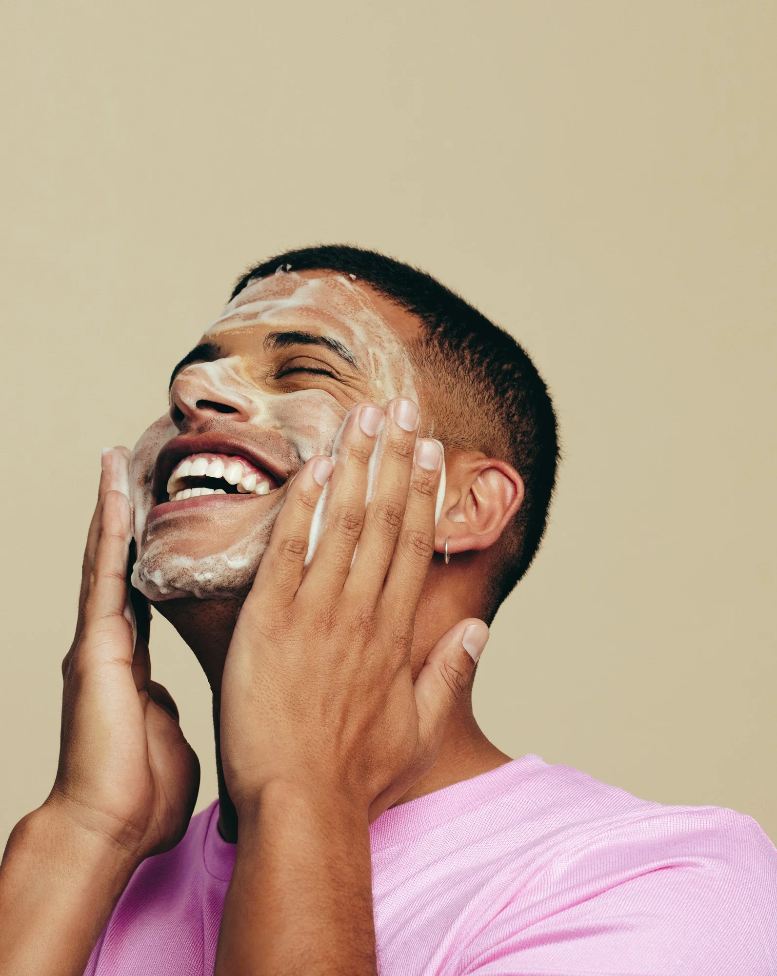 A young man with short black hair applying soap to his face, smiling with eyes closed, wearing a pink shirt against a beige background.