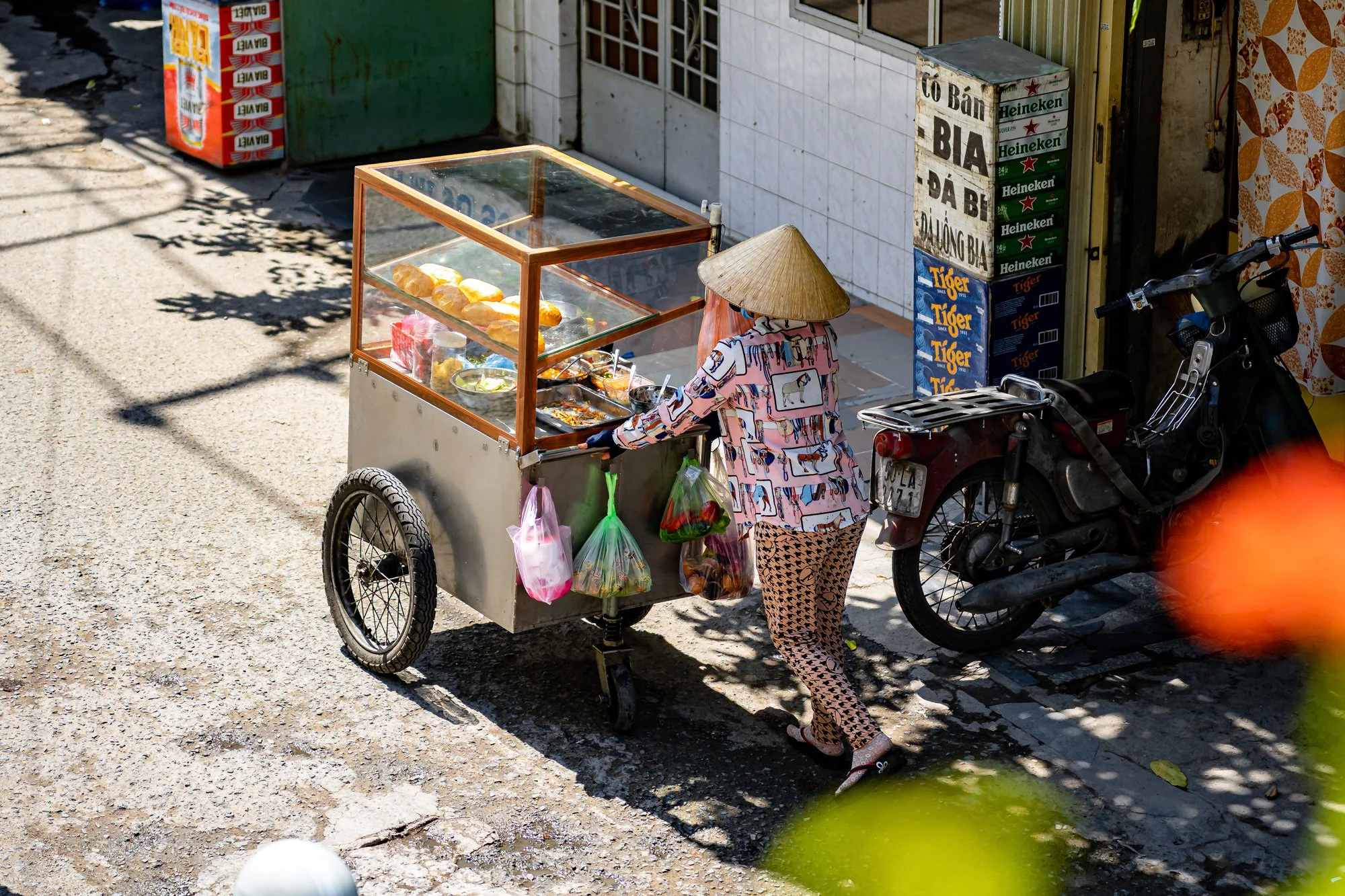 A street vendor with a cart selling food or snacks in an urban area, with a person wearing patterned clothing and a conical hat. The scene includes a motorcycle, stacked boxes, and signage in Vietnamese.