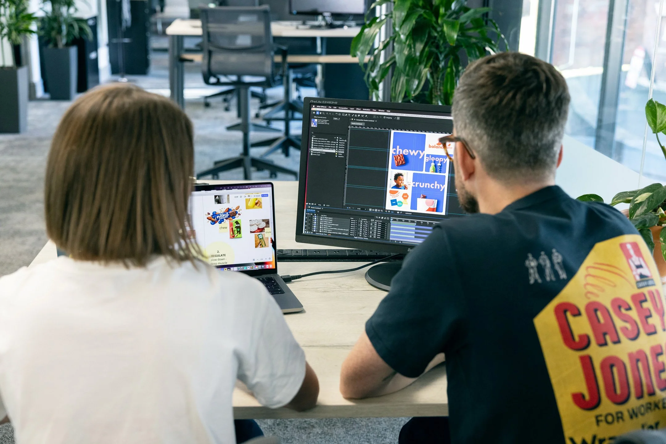 Two people working at a desk with computers in an office, looking at a design on the large screen and a tablet with colorful graphics.