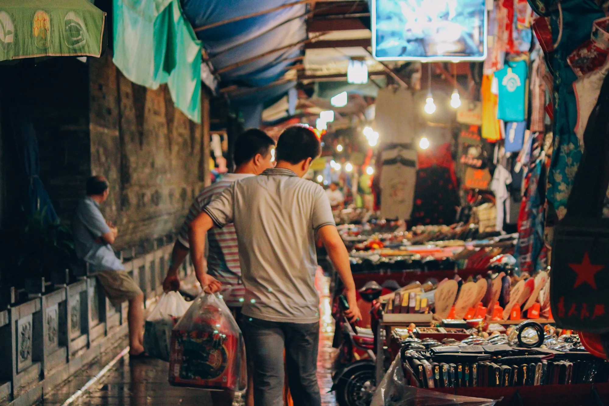 Two young men are shopping at an outdoor market stall that has various items including clothing, shoes, and accessories displayed on tables and racks. The scene is illuminated by warm overhead lights and there are other shoppers in the background.