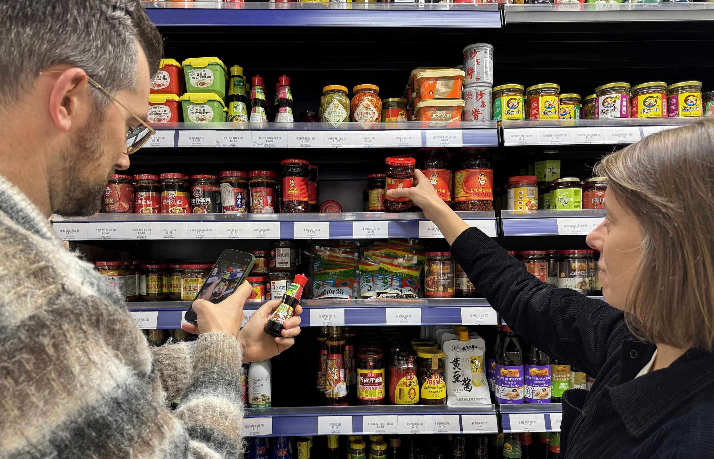 People shopping for Asian sauces and condiments on store shelves, with one person holding a small bottle and another selecting a jar.