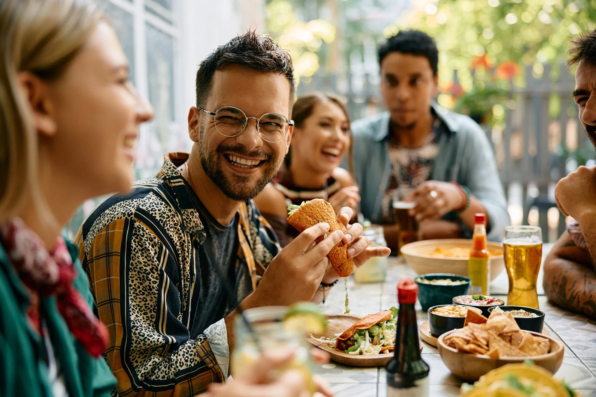 Group of friends at a table enjoying food together outdoors, with a focus on a smiling man holding a sandwich.