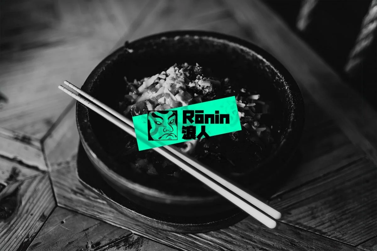 A black bowl of Asian noodles with chopsticks resting on top, placed on a wooden table, with a neon green graphic overlay featuring a stylized face and the text 'Rōnin' and Japanese characters.