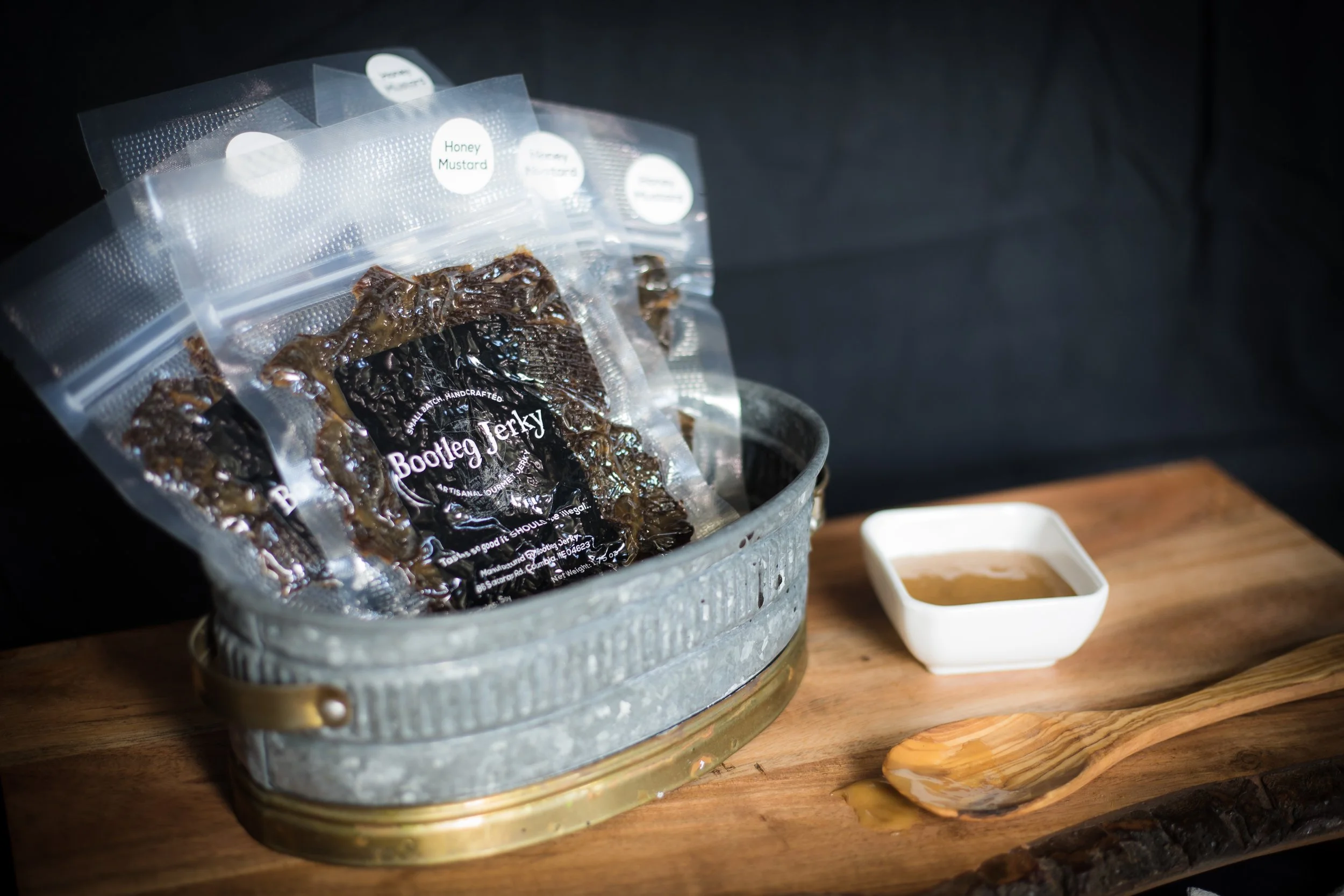 Vacuum sealed packages of sweet and tangy honey mustard beef jerky in an old fashioned steel tub sit beside a dish of mustard sauce.