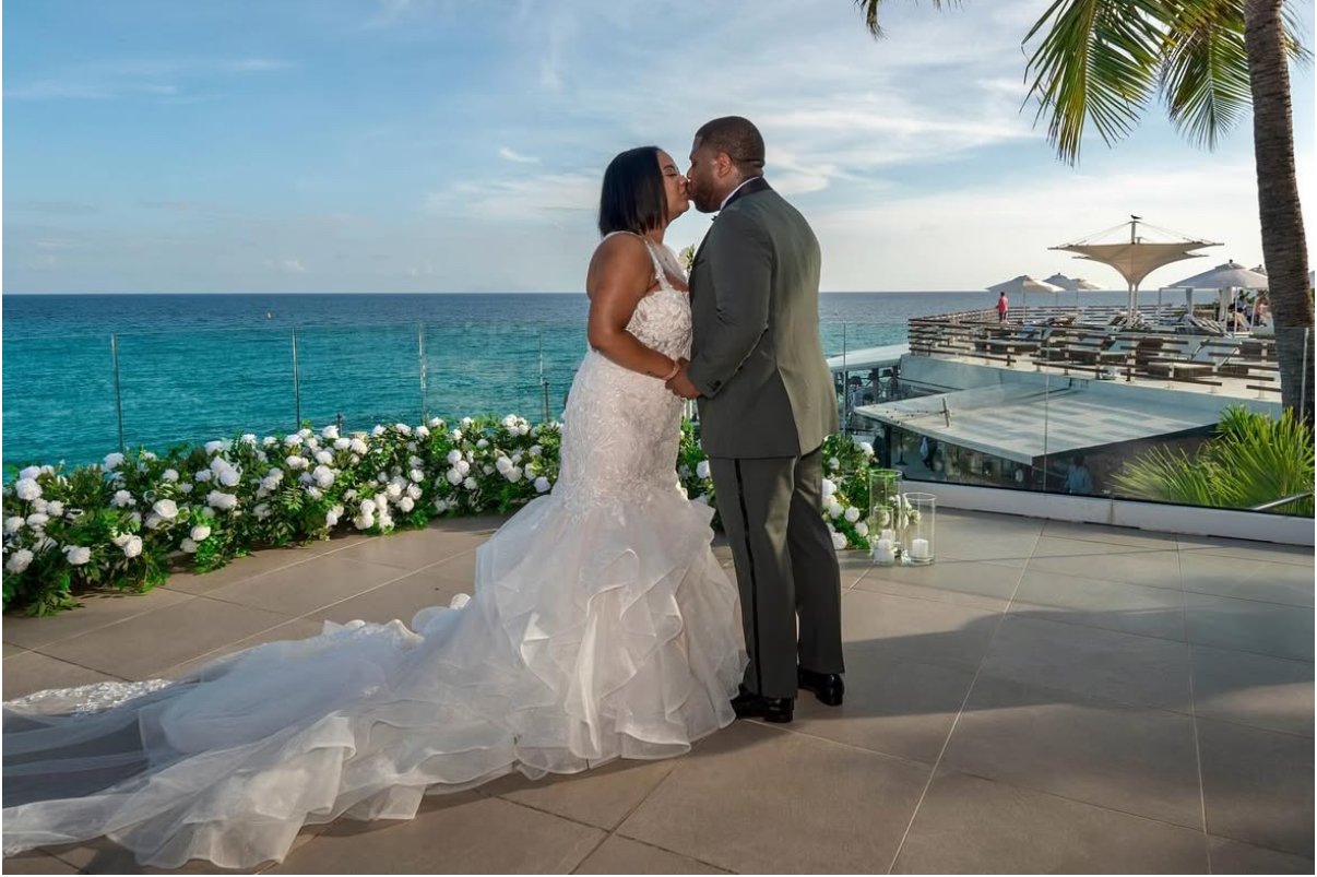 A couple in wedding attire sharing a kiss on a beachside terrace with ocean view, white flowers, and palm trees.