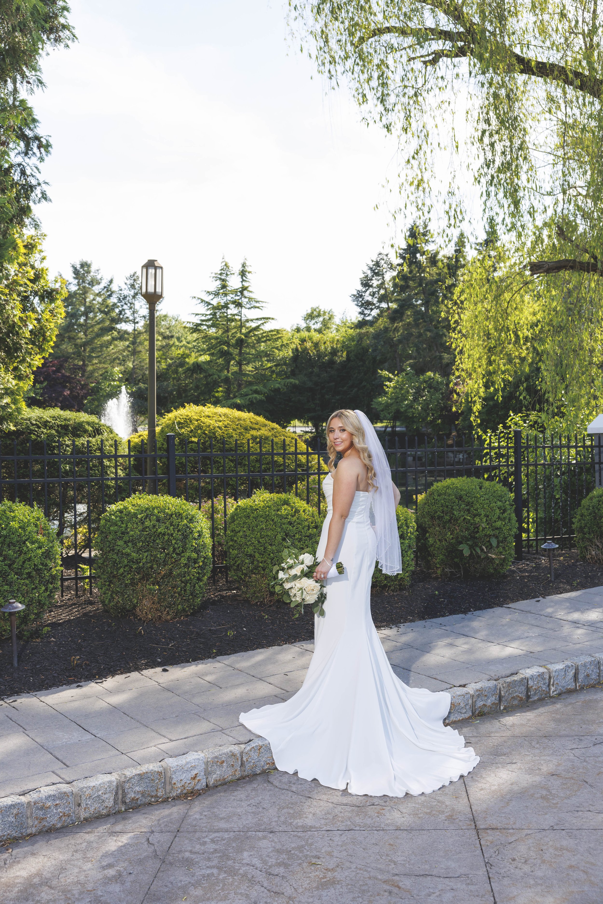 A bride in a white wedding gown holding a bouquet of white roses standing outdoors on a paved pathway, with greenery, bushes, and a black iron fence in the background, under a tree with overhanging branches.