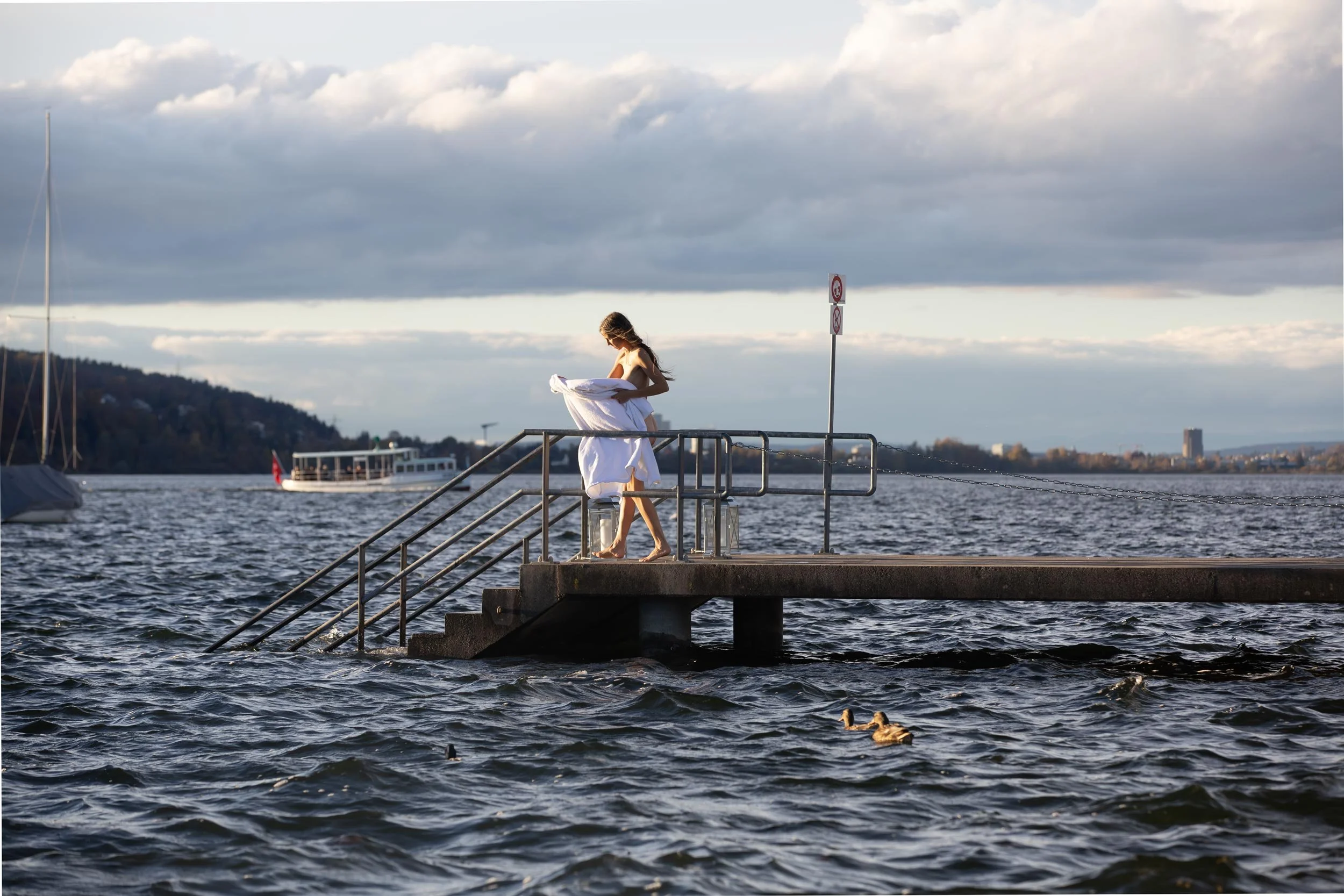 Eine Frau steht auf einer Anlegestelle am Wasser, umgeben von Wasser und Booten, mit einem Hund im Wasser, bei bewölktem Himmel.