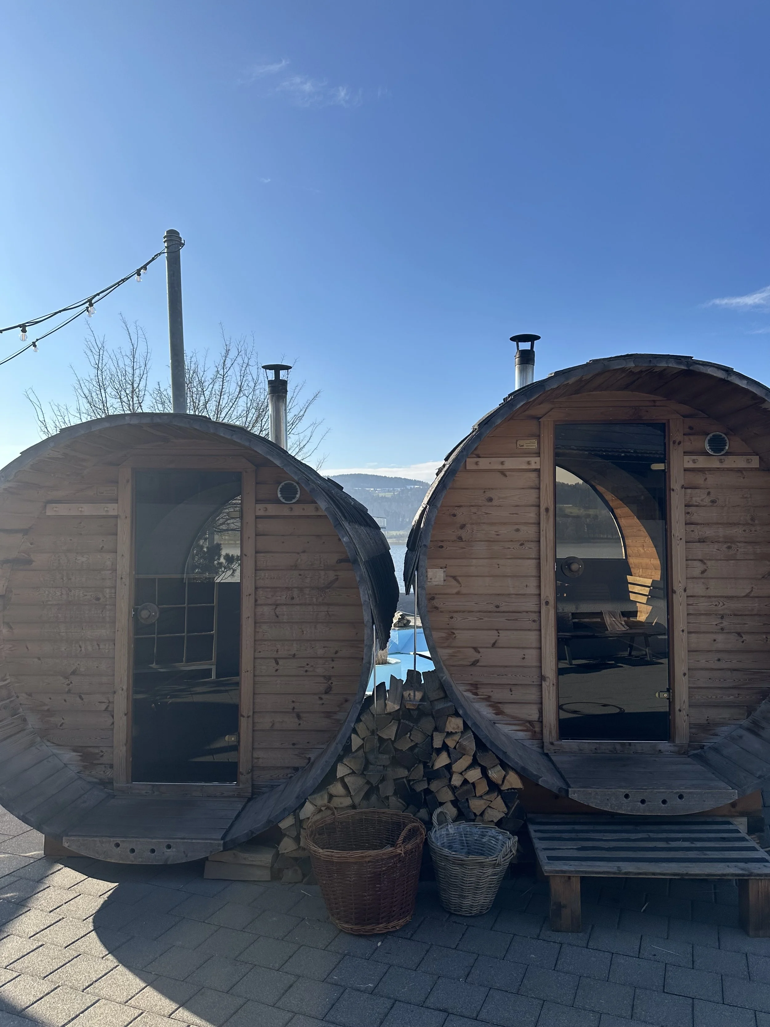 Zwei runde Holzhütten nebeneinander, mit Holzscheiten darunter und geflochtenen Körben davor, im Freien bei sonnigem Himmel.