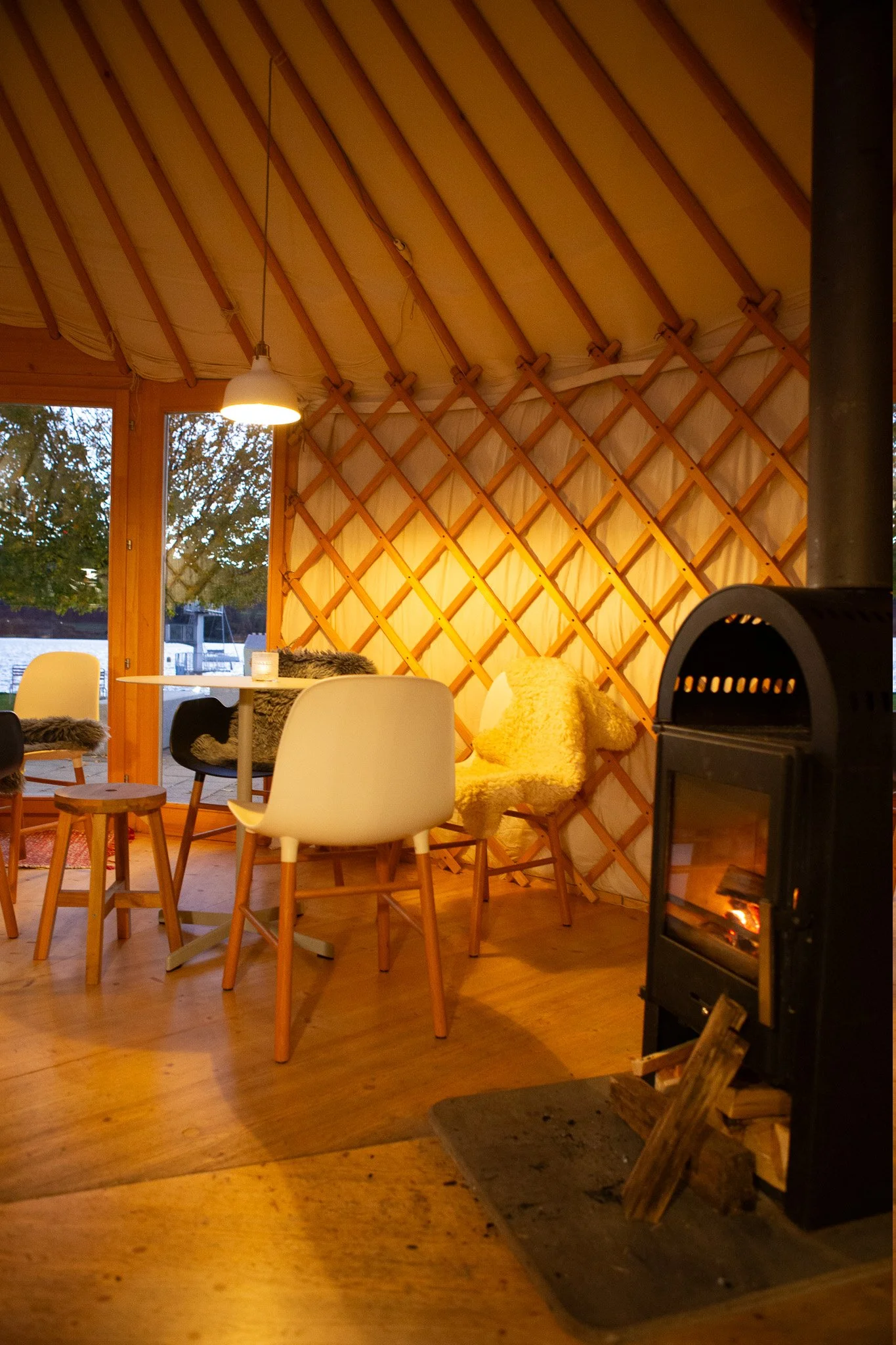 Gemütliches Innere eines Zeltes mit Holzmöbeln, einem Kamin und einer Lichterkette, mit Blick nach draußen auf Wasser und Bäume.