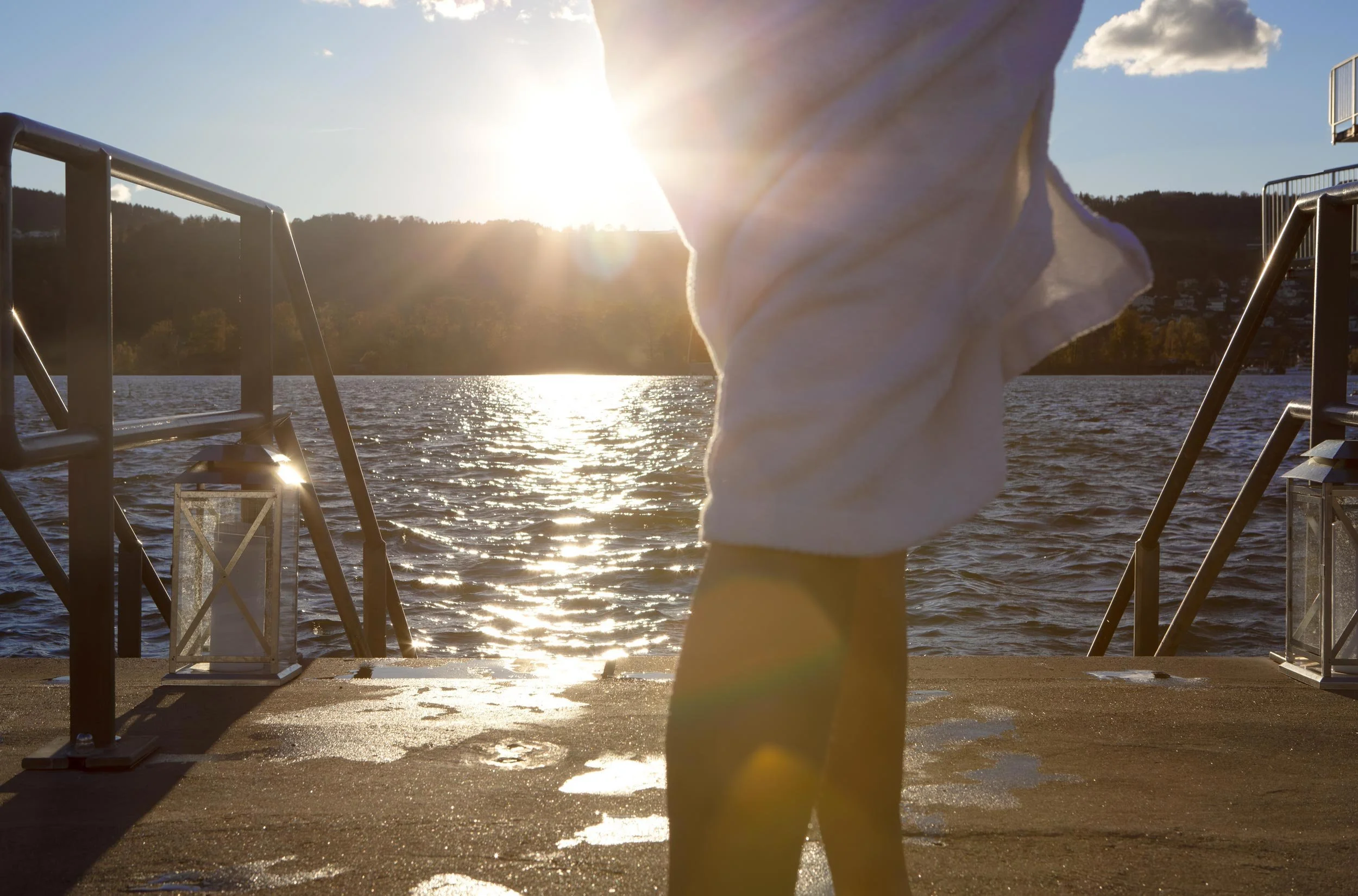Person steht auf einem Bootssteg bei Sonnenuntergang, Wasser im Hintergrund, Sonnenlicht reflektiert auf dem Wasser.