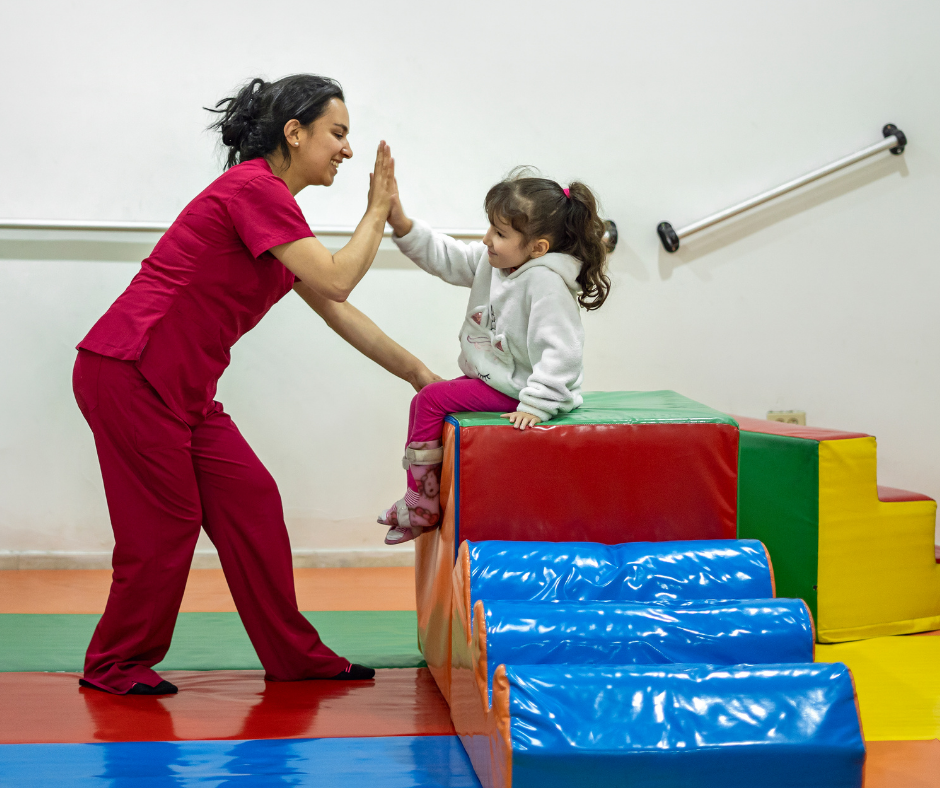 A pediatric occupational therapist is wear magenta scrubs and is giving a child a high five. The child is sitting at the top of a soft play climber.