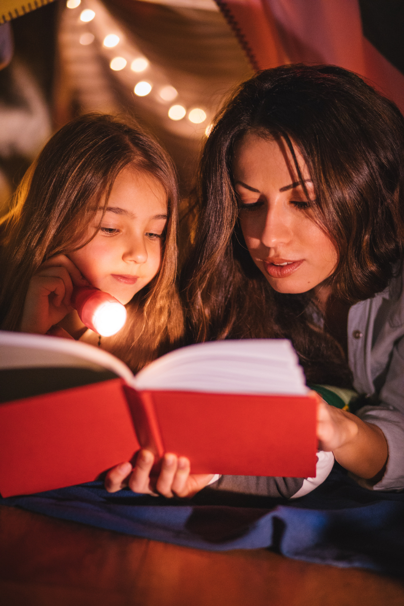 Mom and child reading a book together at bedtime, in dim lighting, by flashlight. Both look peaceful, calm and content