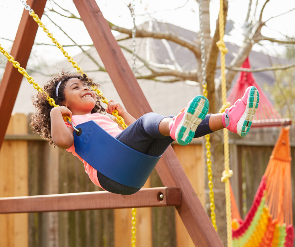A Black child swings joyfully on a backyard swing set in the afternoon sun, capturing the kind of active, sensory-rich outdoor movement that helps neurodivergent children regulate and sleep better at night.