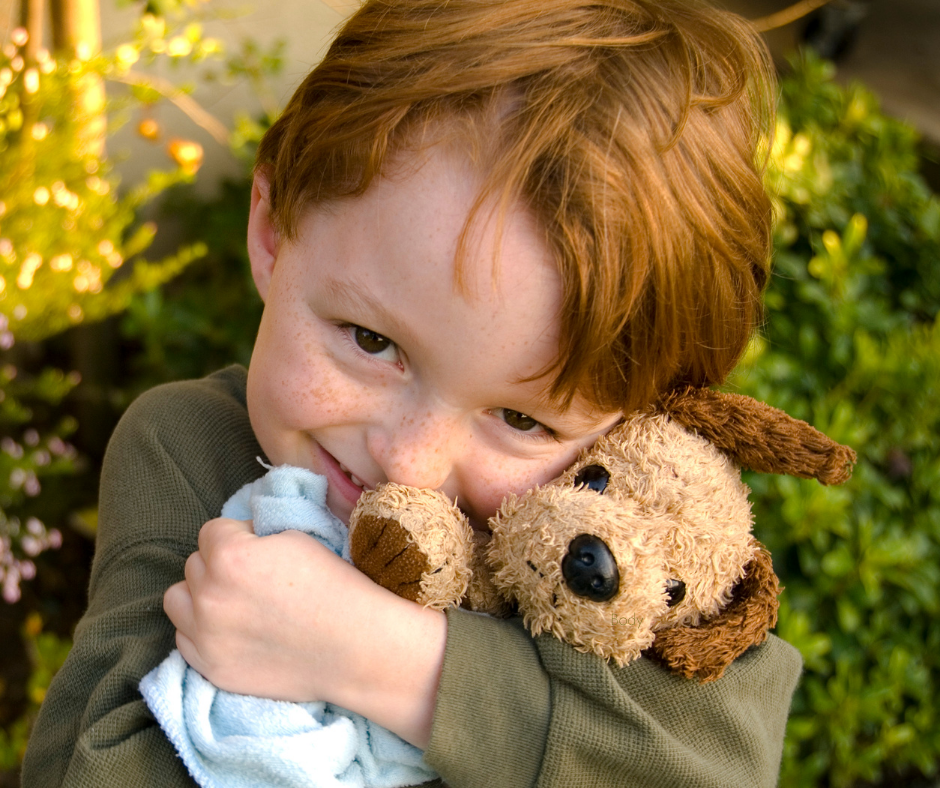 A red-haired child with freckles holds a well-loved teddy bear and blanket outdoors in natural light, capturing the comfort objects and sensory needs that are part of many neurodivergent children's sleep lives.