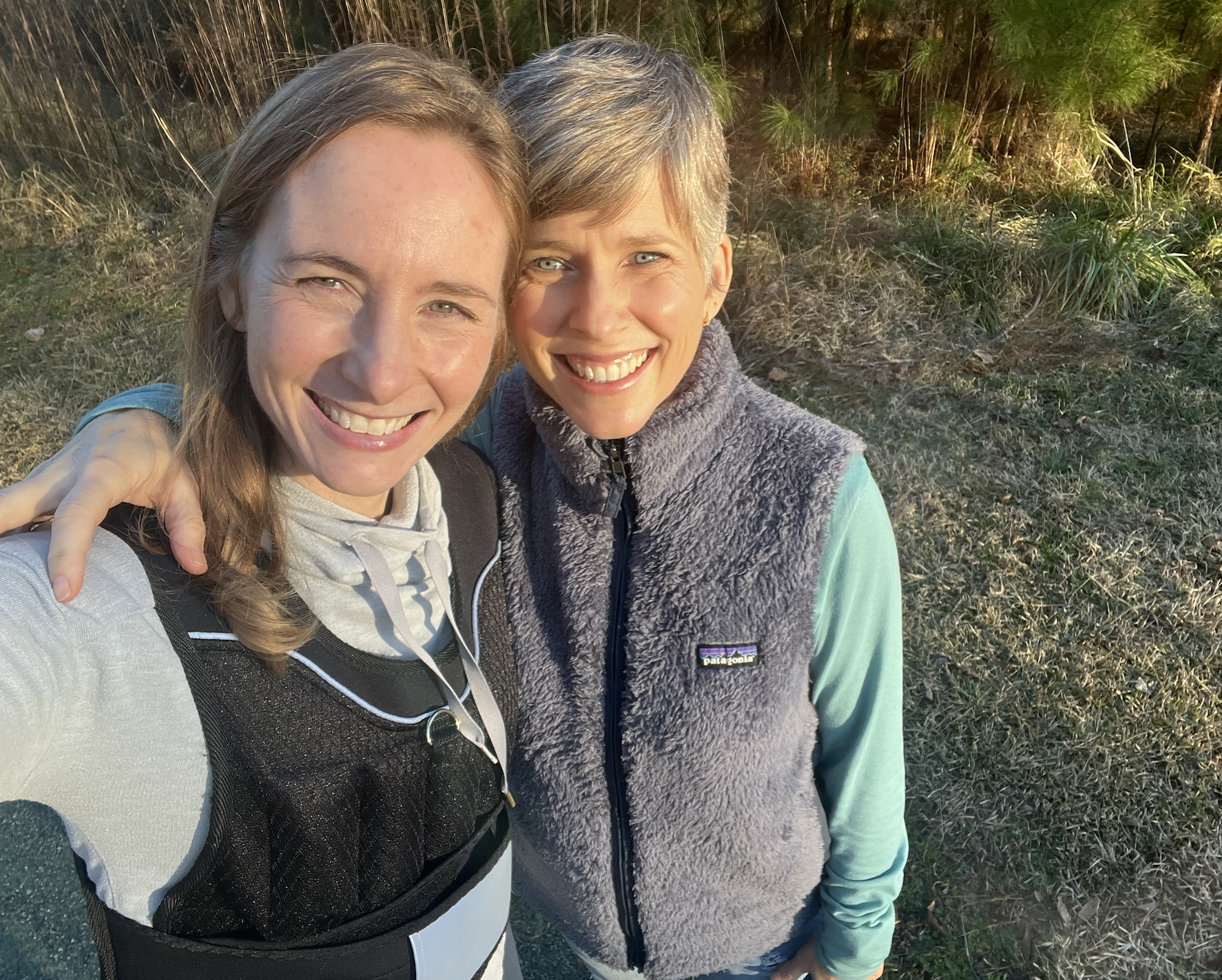 Two women in perimenopause, one wearing a weighted vest, with their arms around each other and smiling at the camera. They are at a park and have been walking.